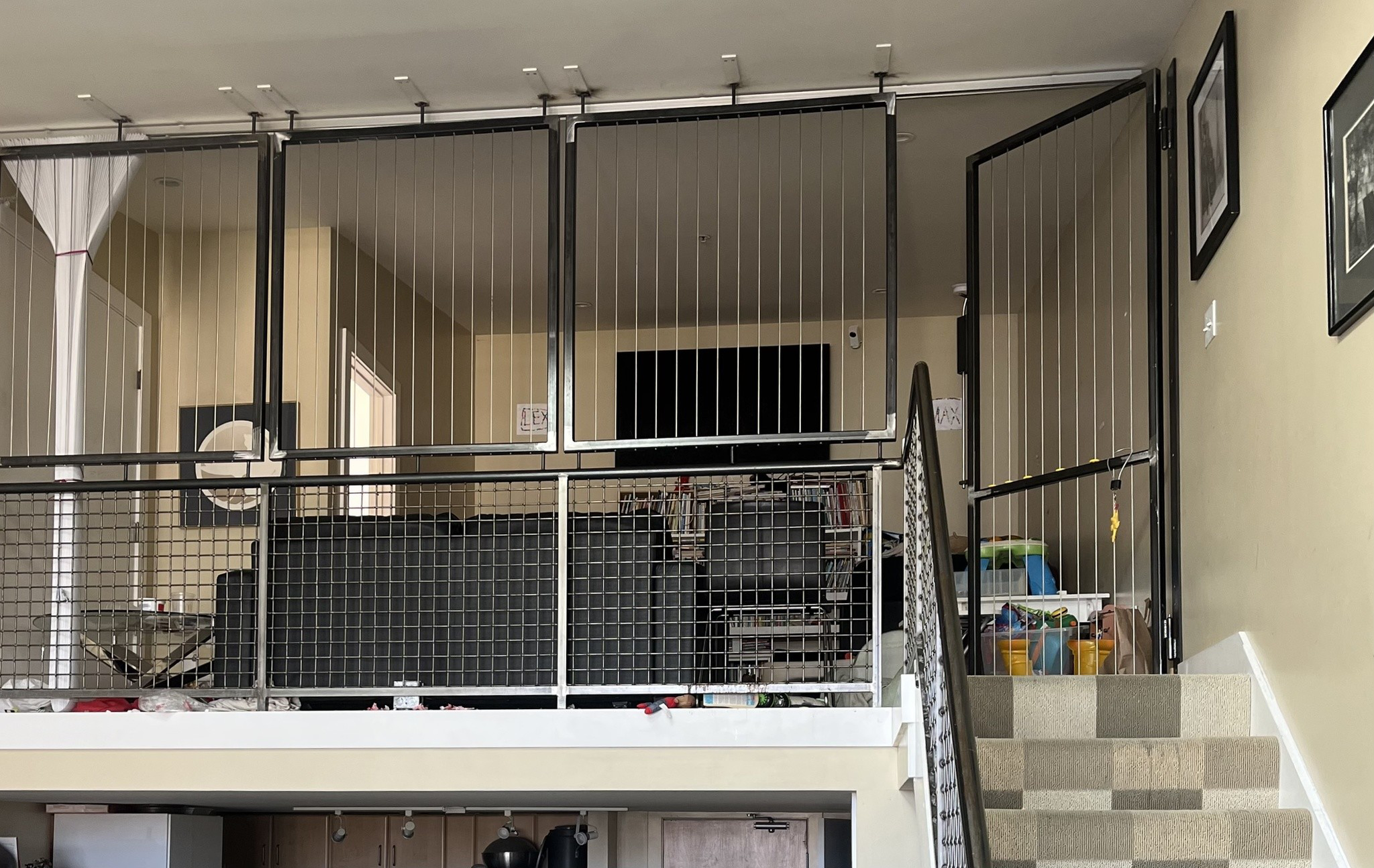 A loft area with black wire safety gates surrounds a living space with a TV and furniture, a staircase with patterned carpet leads up to it.