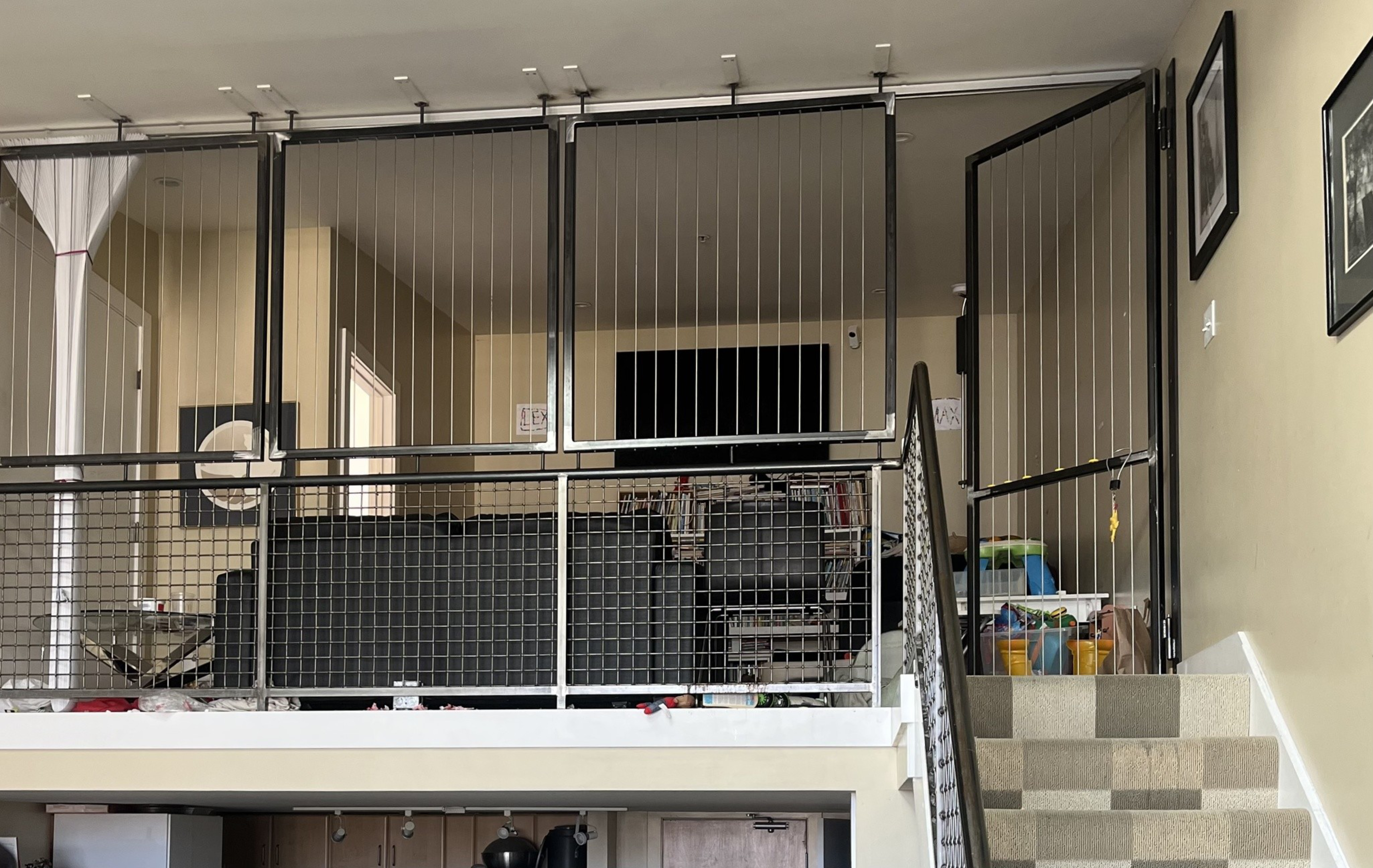 A loft area with black wire safety gates surrounds a living space with a TV and furniture, a staircase with patterned carpet leads up to it.