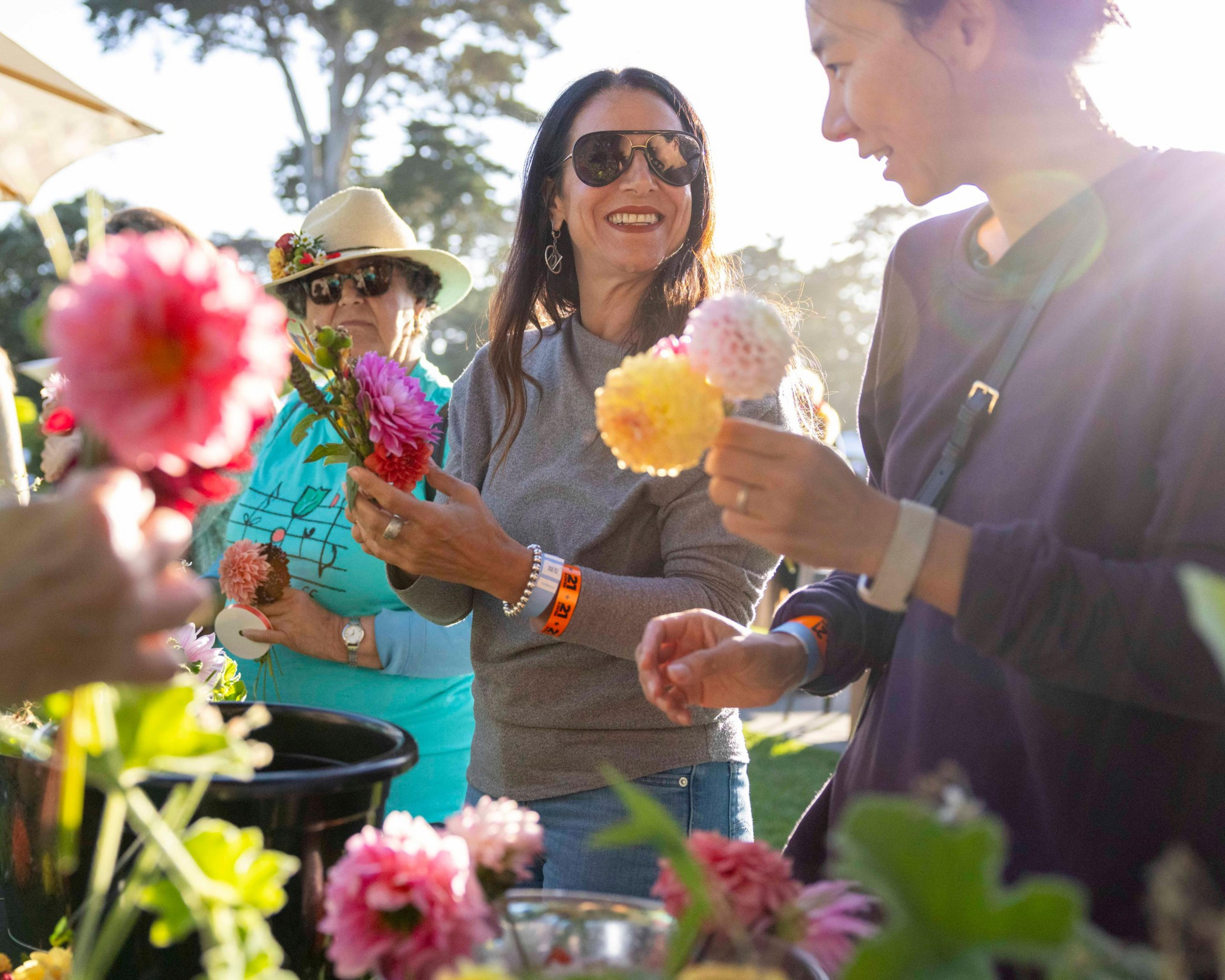 Three women are outdoors, smiling and holding vibrant flowers, with trees visible in the background. They are enjoying a sunny day, wearing casual clothes and glasses.