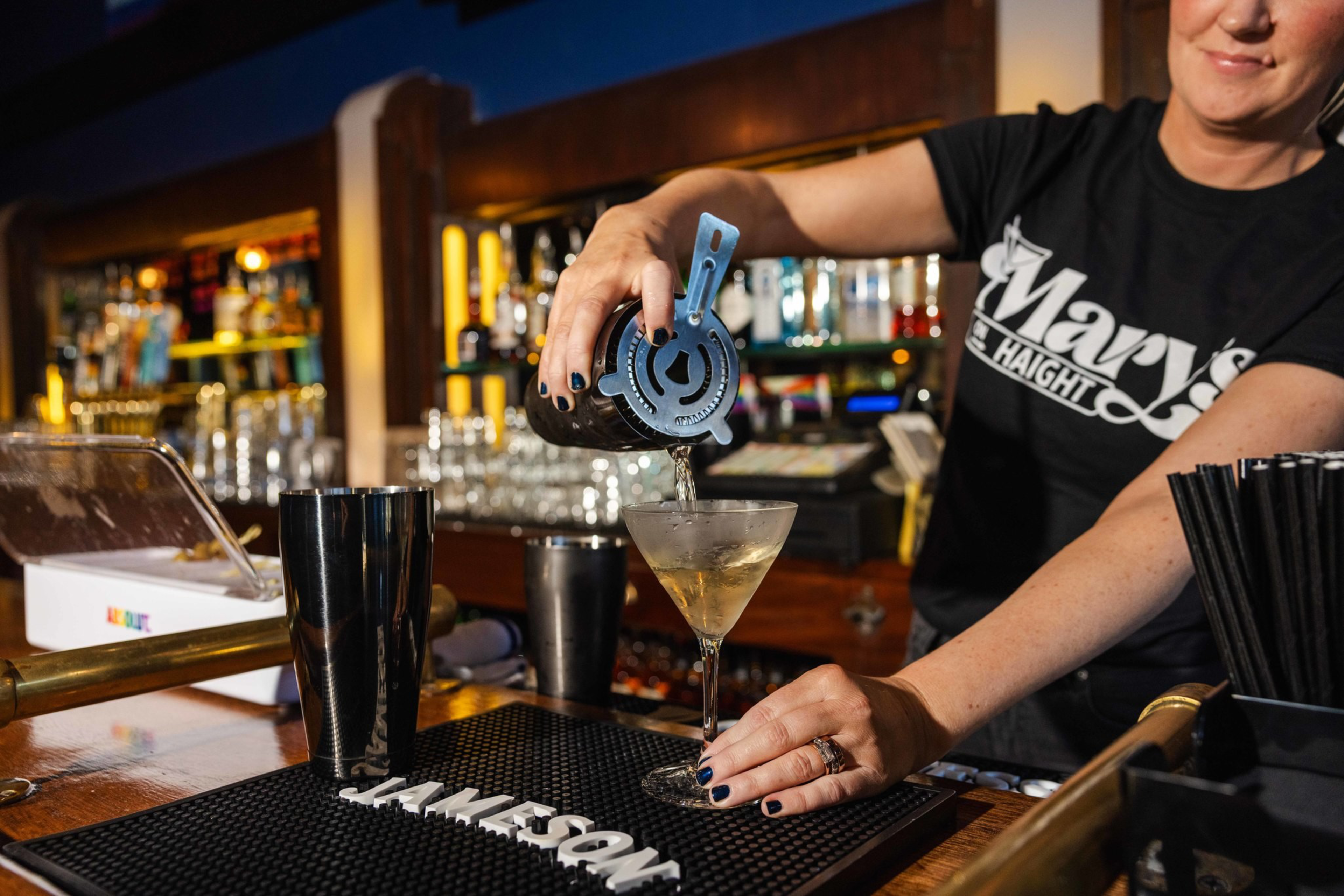 A bartender with black nail polish pours a cocktail into a martini glass using a strainer at a bar with glasses and bottles in the background.