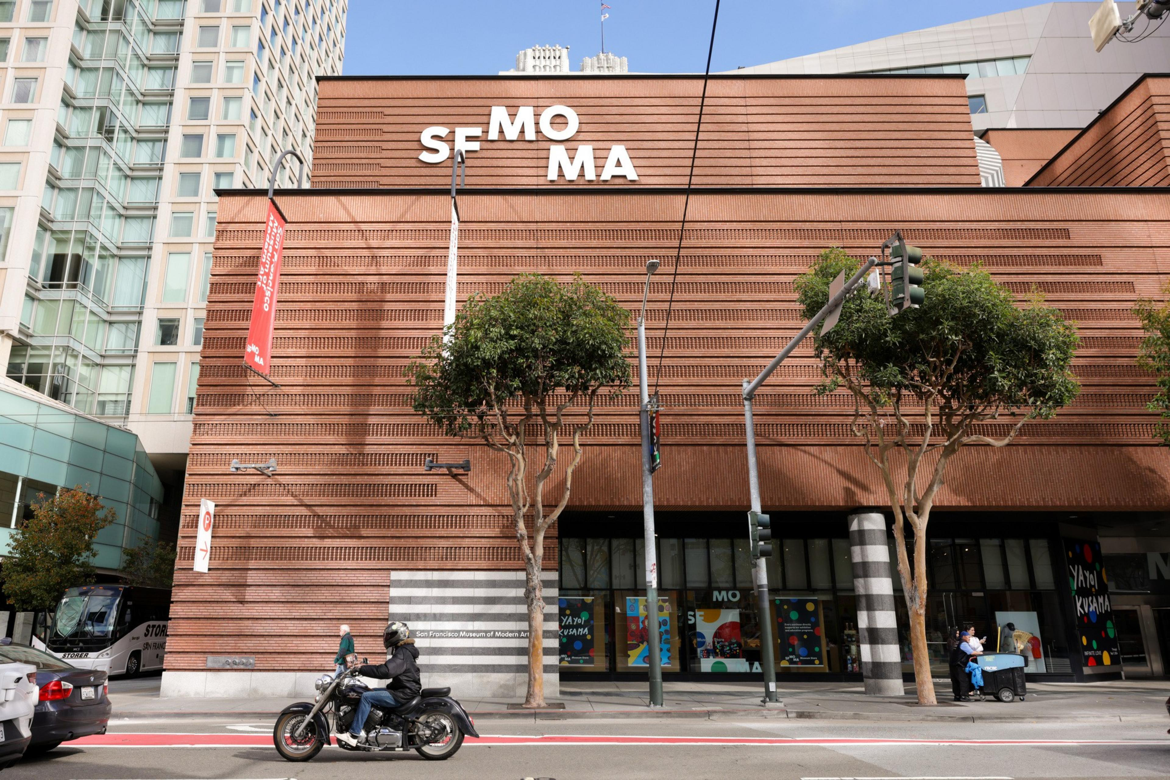 A museum stands behind trees and a motorcyle on the street.