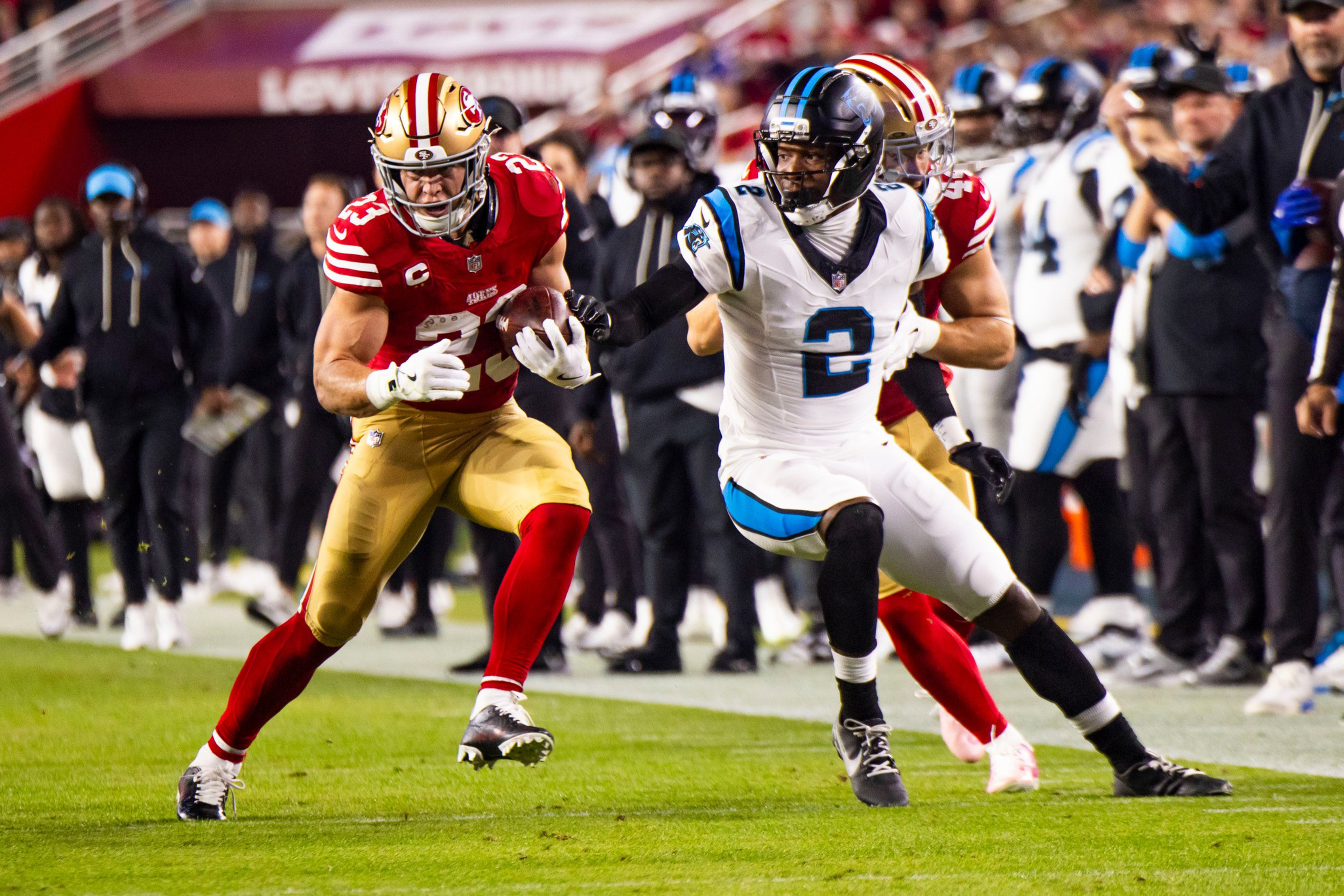 A football player in a red and gold uniform carries the ball while a player in a white and blue uniform tries to tackle him during a game.