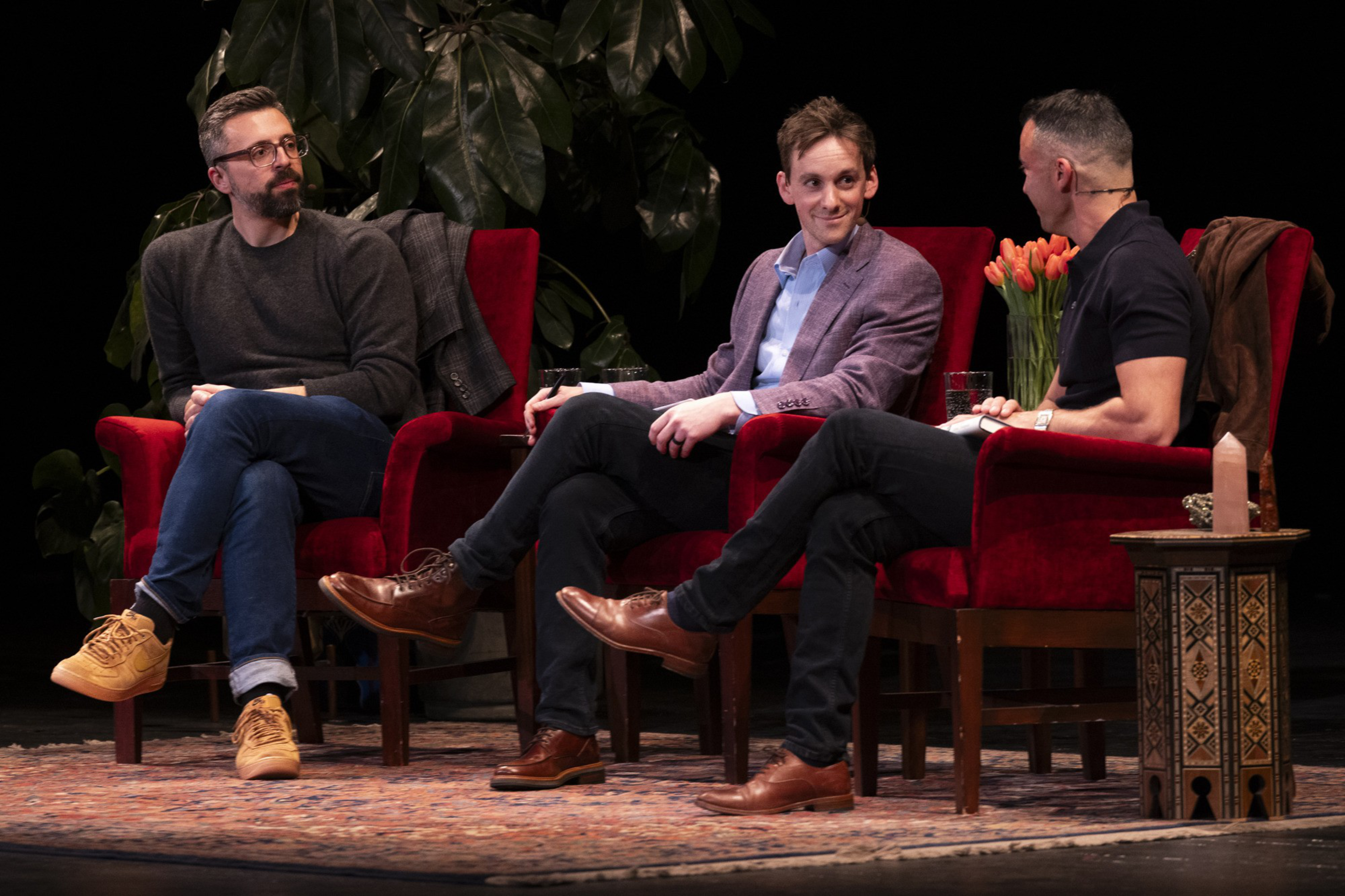 Three men sit in red chairs on stage, engaged in conversation. One holds a notebook. There are plants, tulips, and a patterned table beside them.