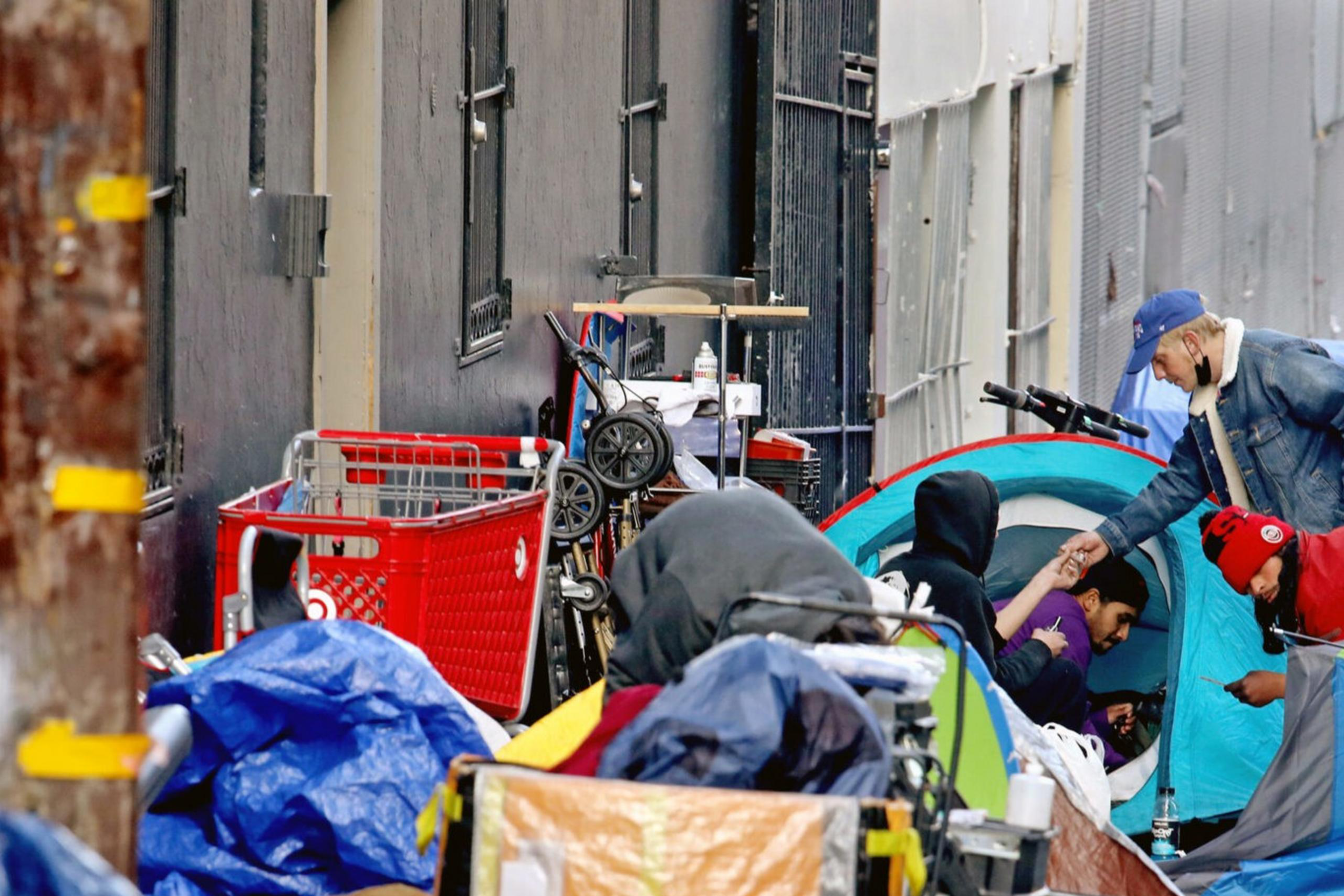 Several people are gathered in a narrow alley cluttered with tents, tarps, and personal belongings. Some are interacting, while others are using phones amidst scattered debris.