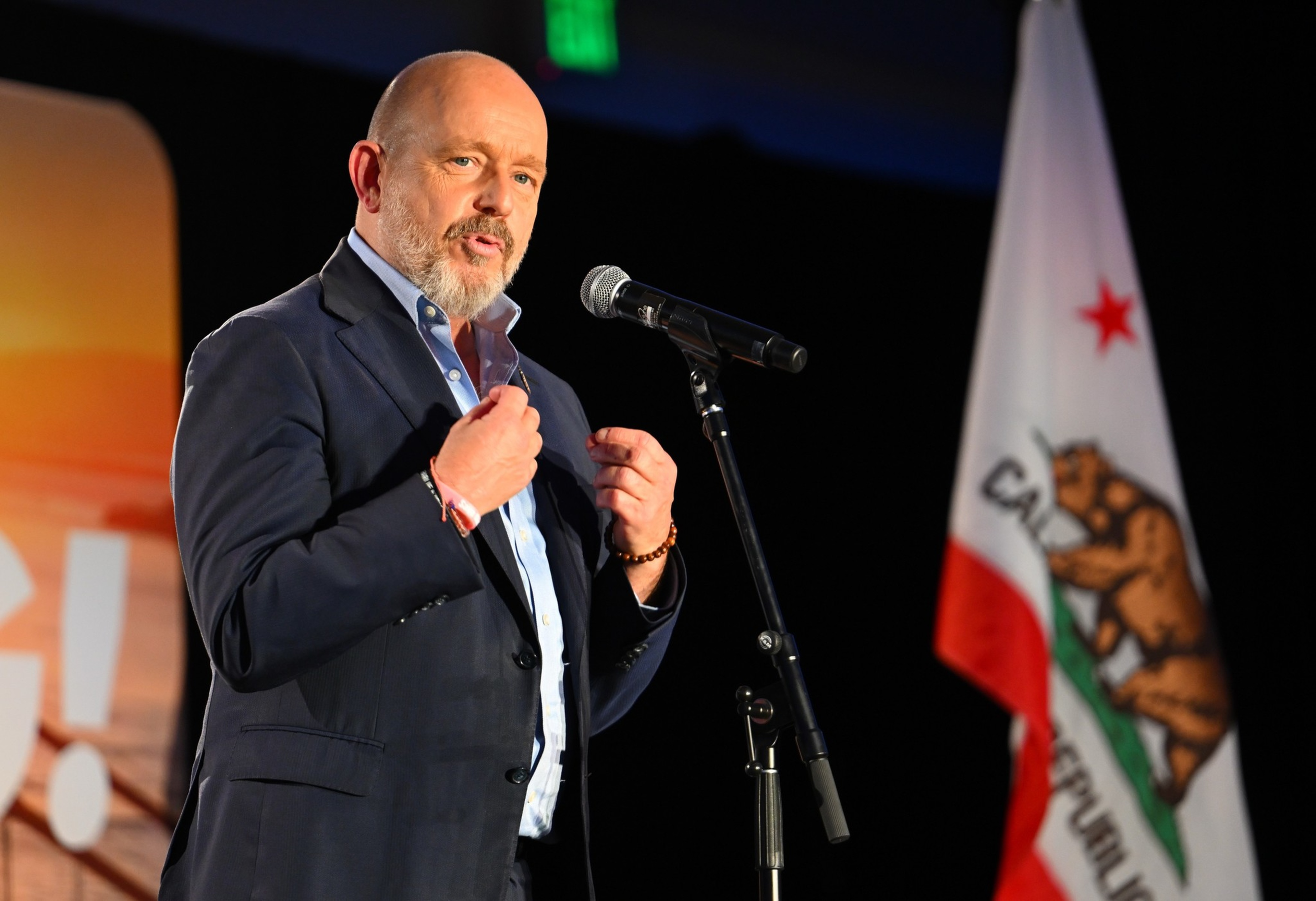 A man in a suit speaks into a microphone with a California state flag visible in the background.