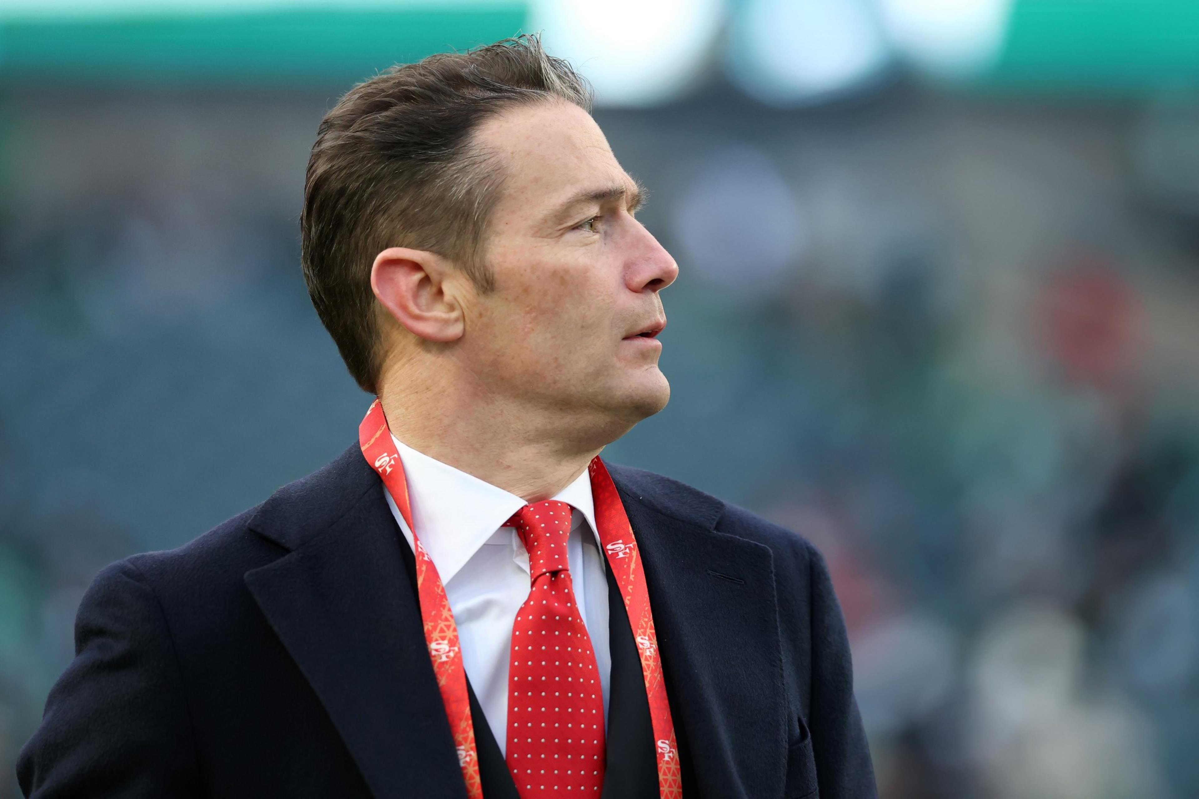 A man wearing a dark suit, white shirt, and red tie looks to the side with a red lanyard around his neck, standing outdoors with blurred background.