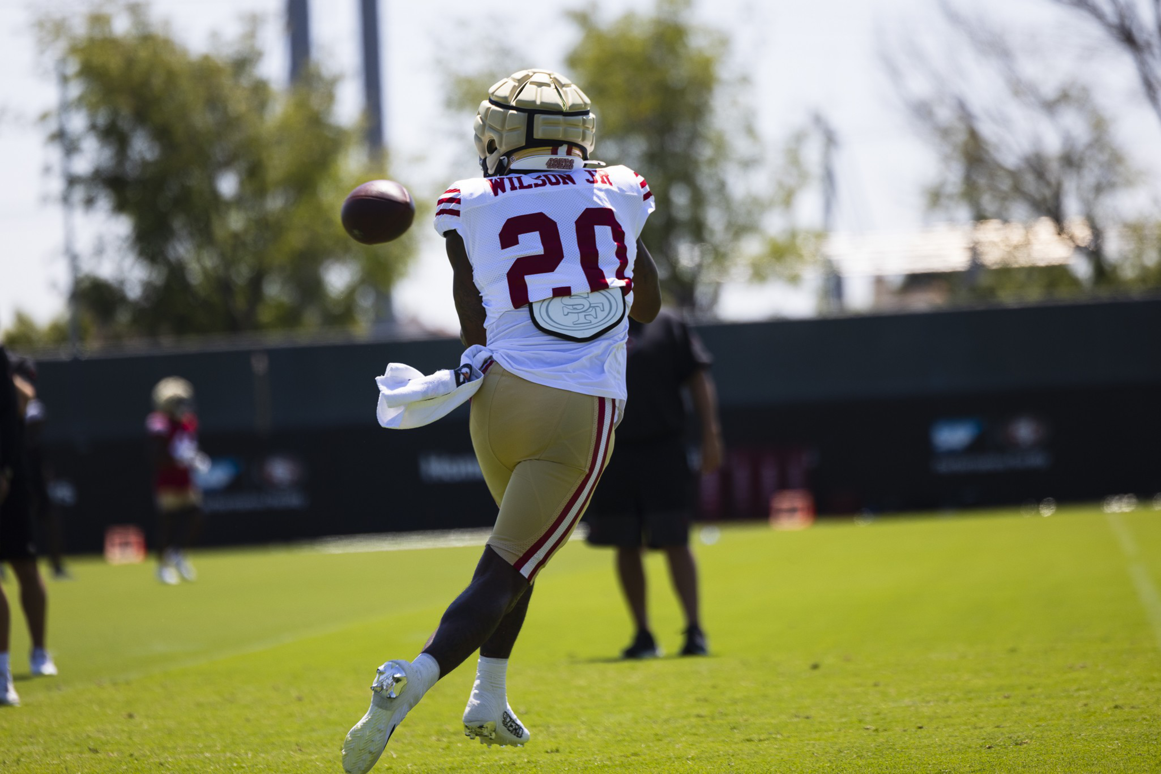 A football player wearing a number 20 jersey catches a ball mid-air on a sunny practice field with trees and other players in the background.