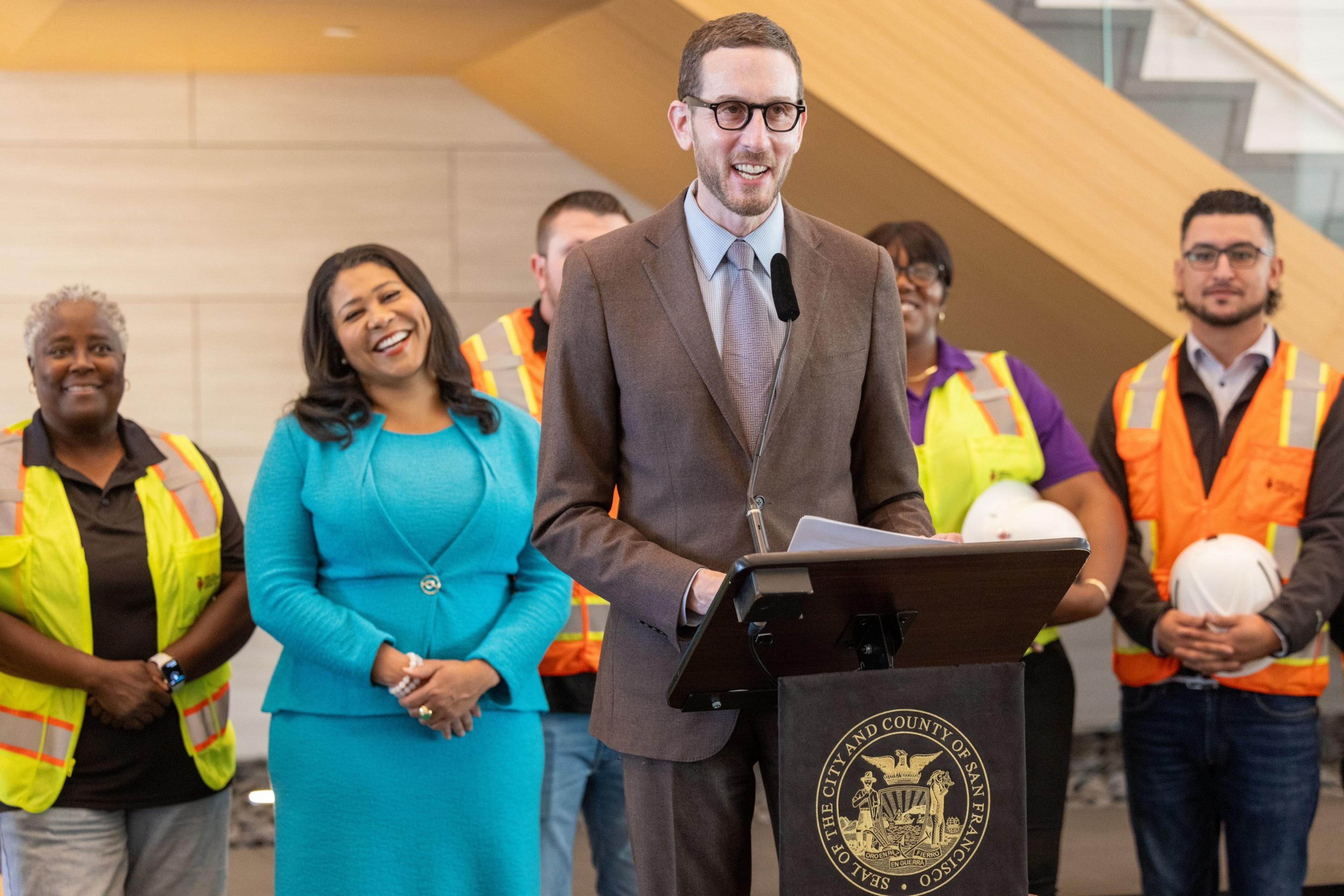 A man in a suit speaks at a podium, flanked by people in safety vests and a woman in a blue outfit, all smiling. They stand in a modern, bright space.