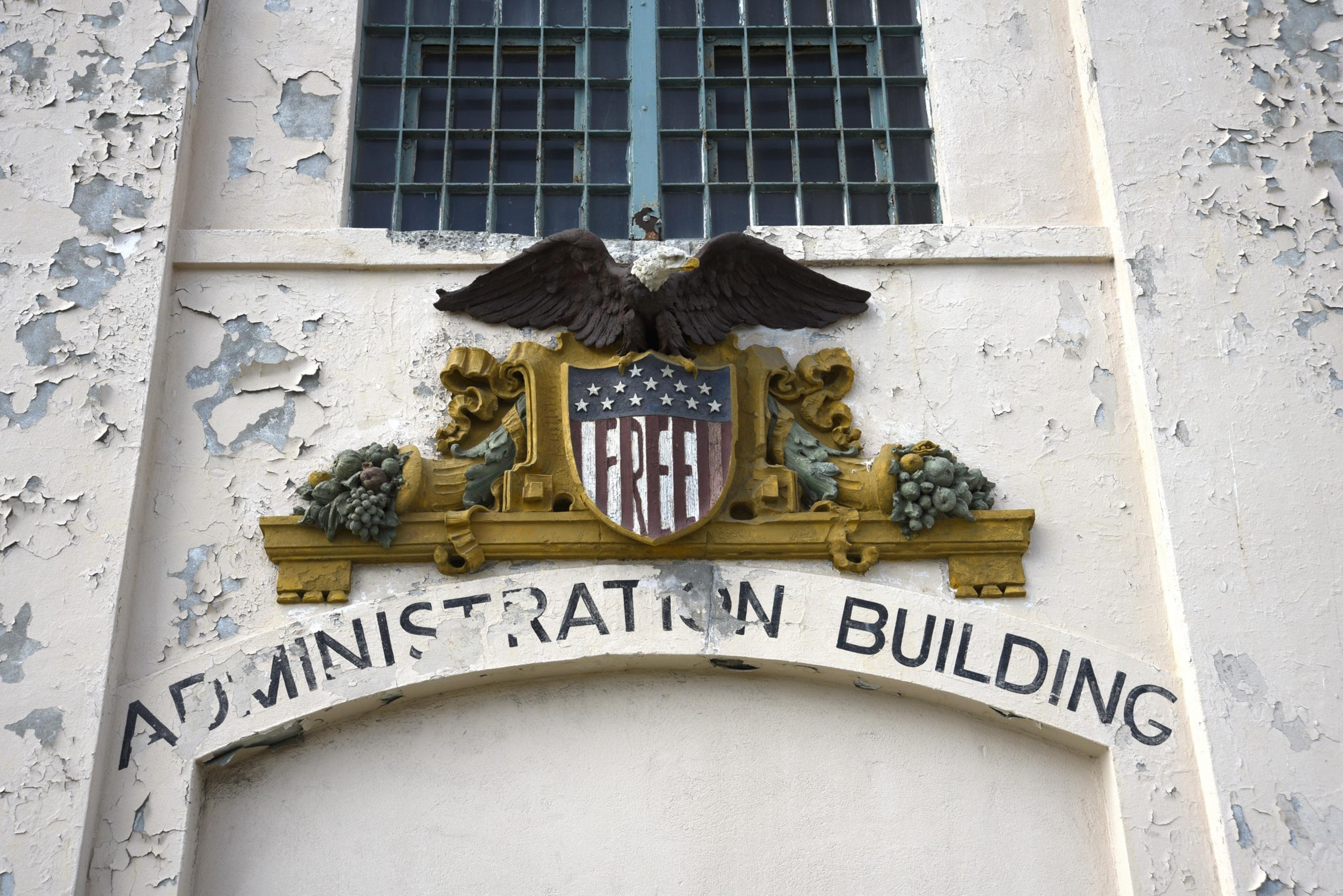 An old building facade features a peeling paint surface, an eagle atop a shield with &quot;FREE,&quot; and an arch labeled &quot;ADMINISTRATION BUILDING&quot; in bold text.