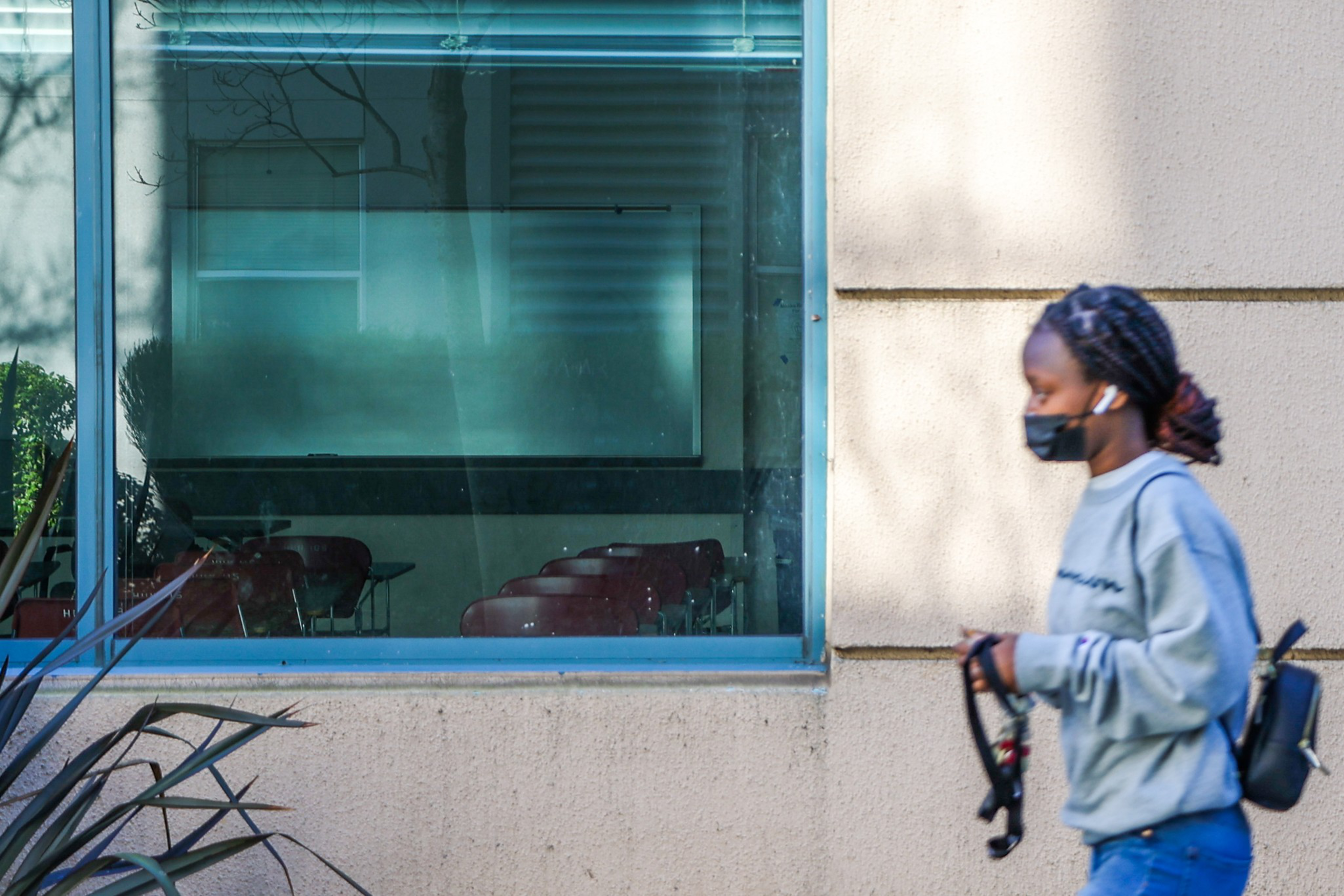 A person wearing a mask and earbuds walks past a beige wall with a large window that shows empty chairs inside a room.