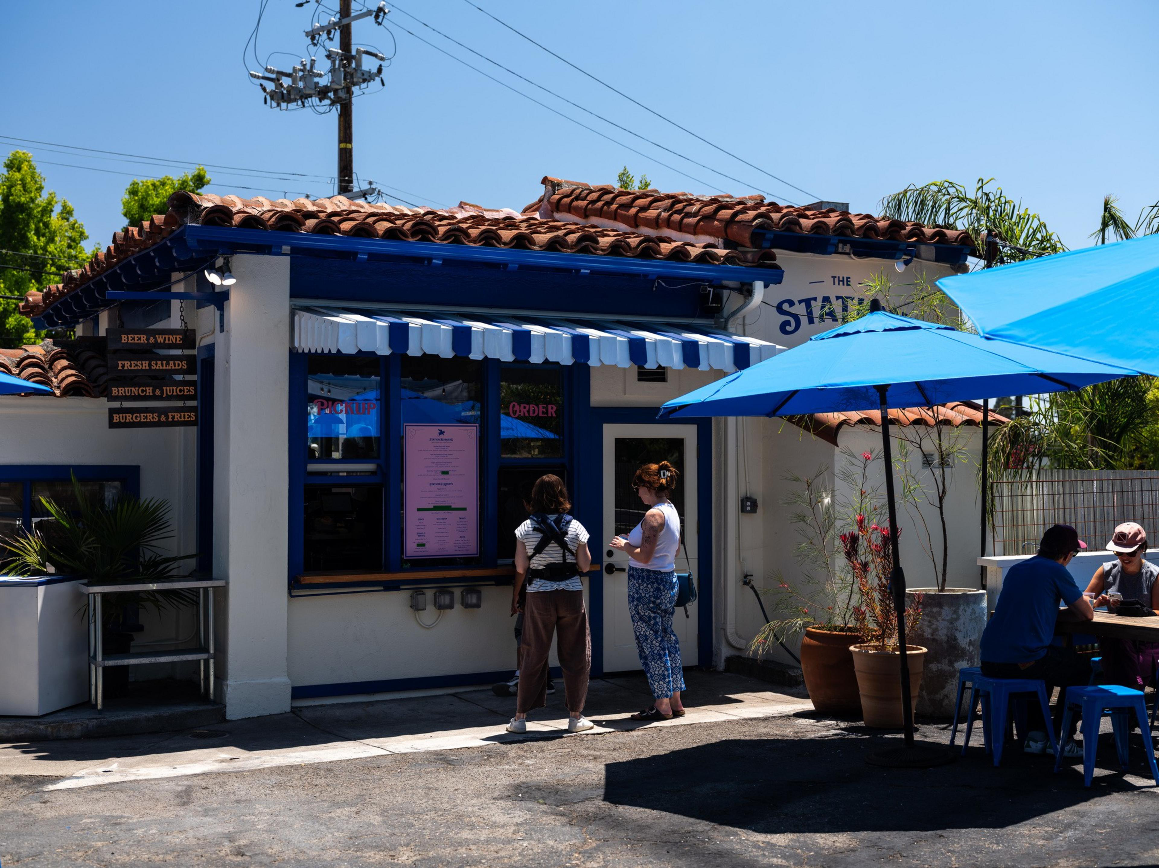 A small eatery with a blue and white awning and signs for beer, brunch, and burgers. Two people stand by the order window, while two others sit under blue umbrellas.