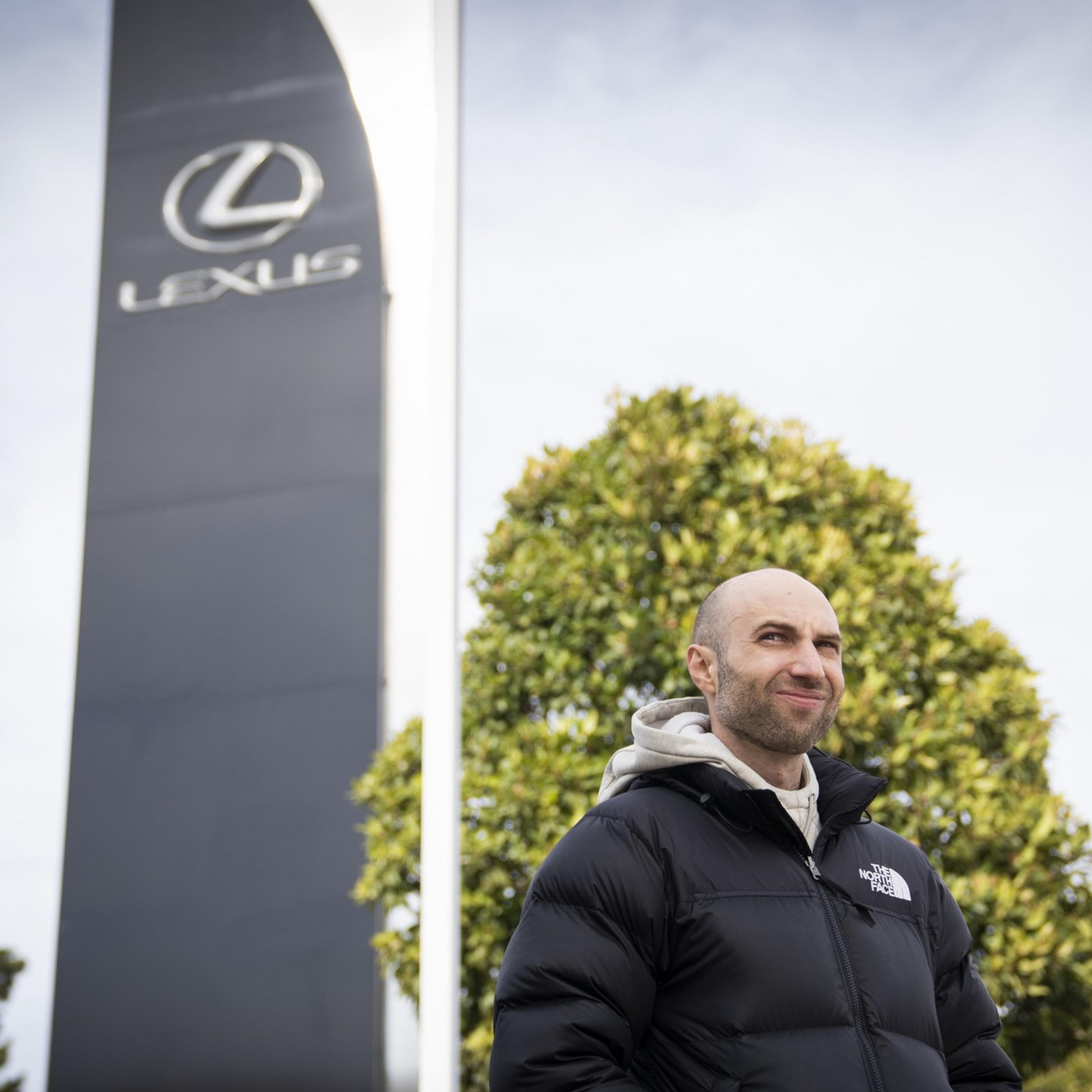 A person wearing a black puffer jacket stands outdoors near a large sign with the Lexus logo, against a backdrop of trees and a cloudy sky.