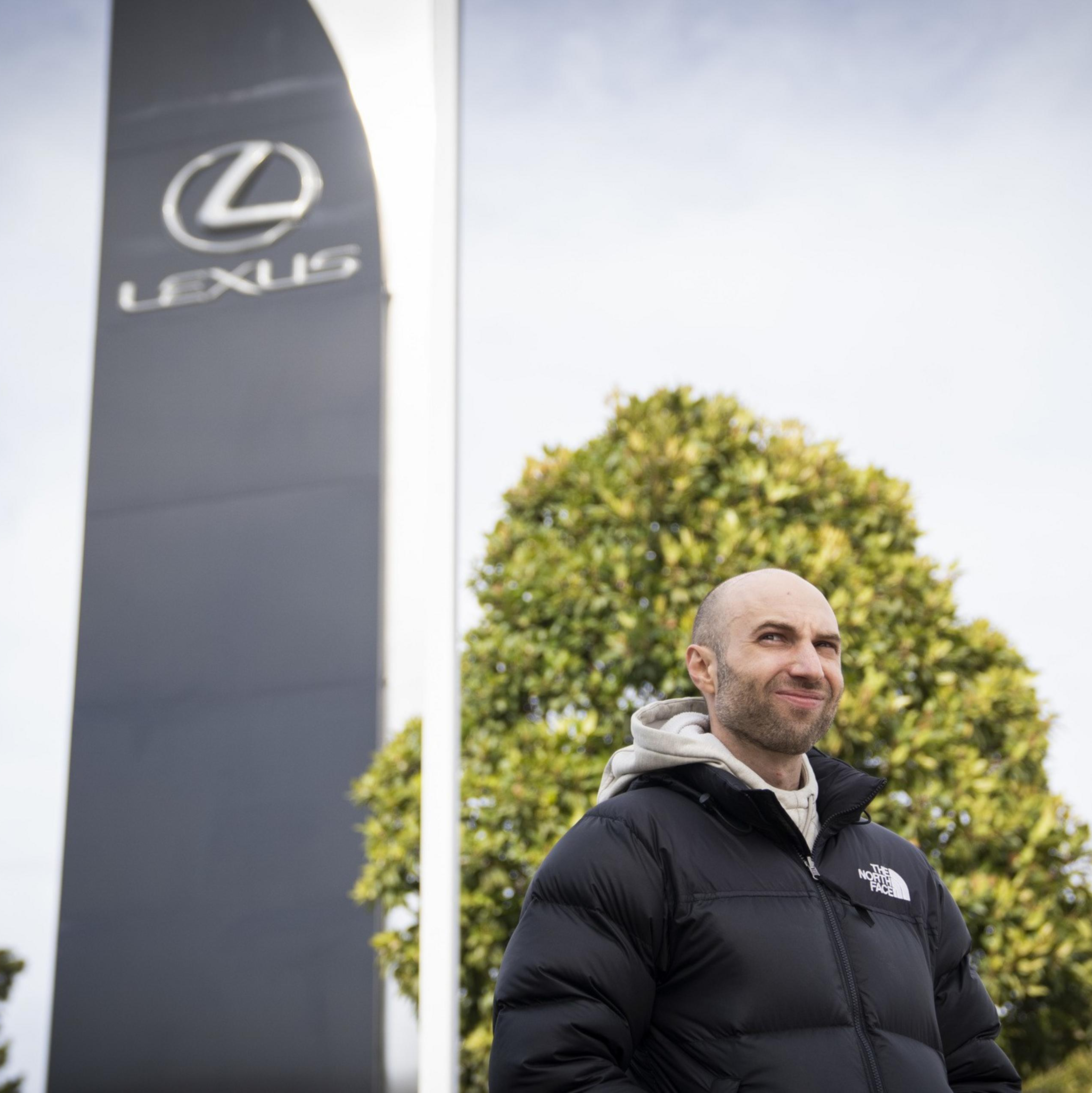 A person wearing a black puffer jacket stands outdoors near a large sign with the Lexus logo, against a backdrop of trees and a cloudy sky.