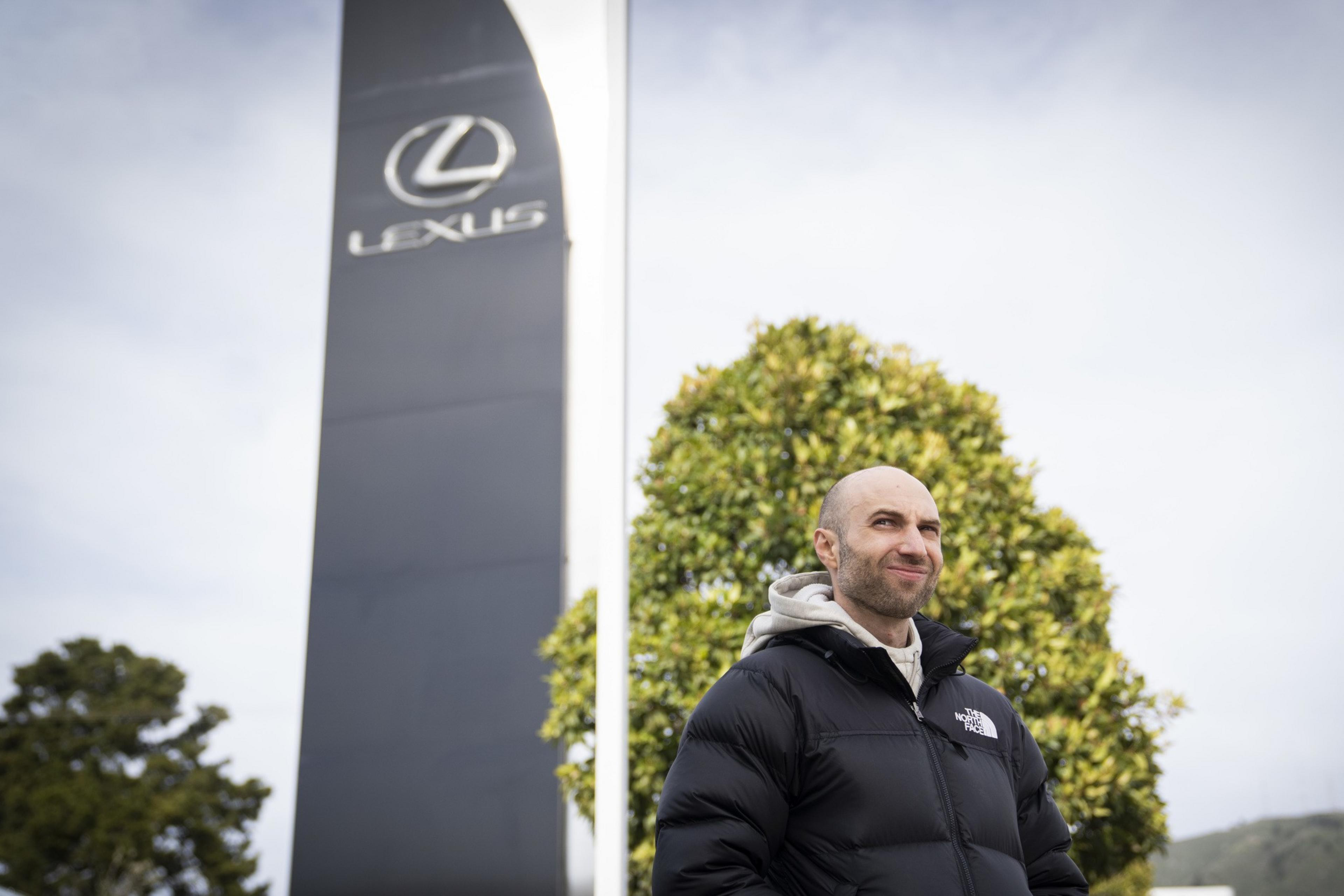 A person wearing a black puffer jacket stands outdoors near a large sign with the Lexus logo, against a backdrop of trees and a cloudy sky.