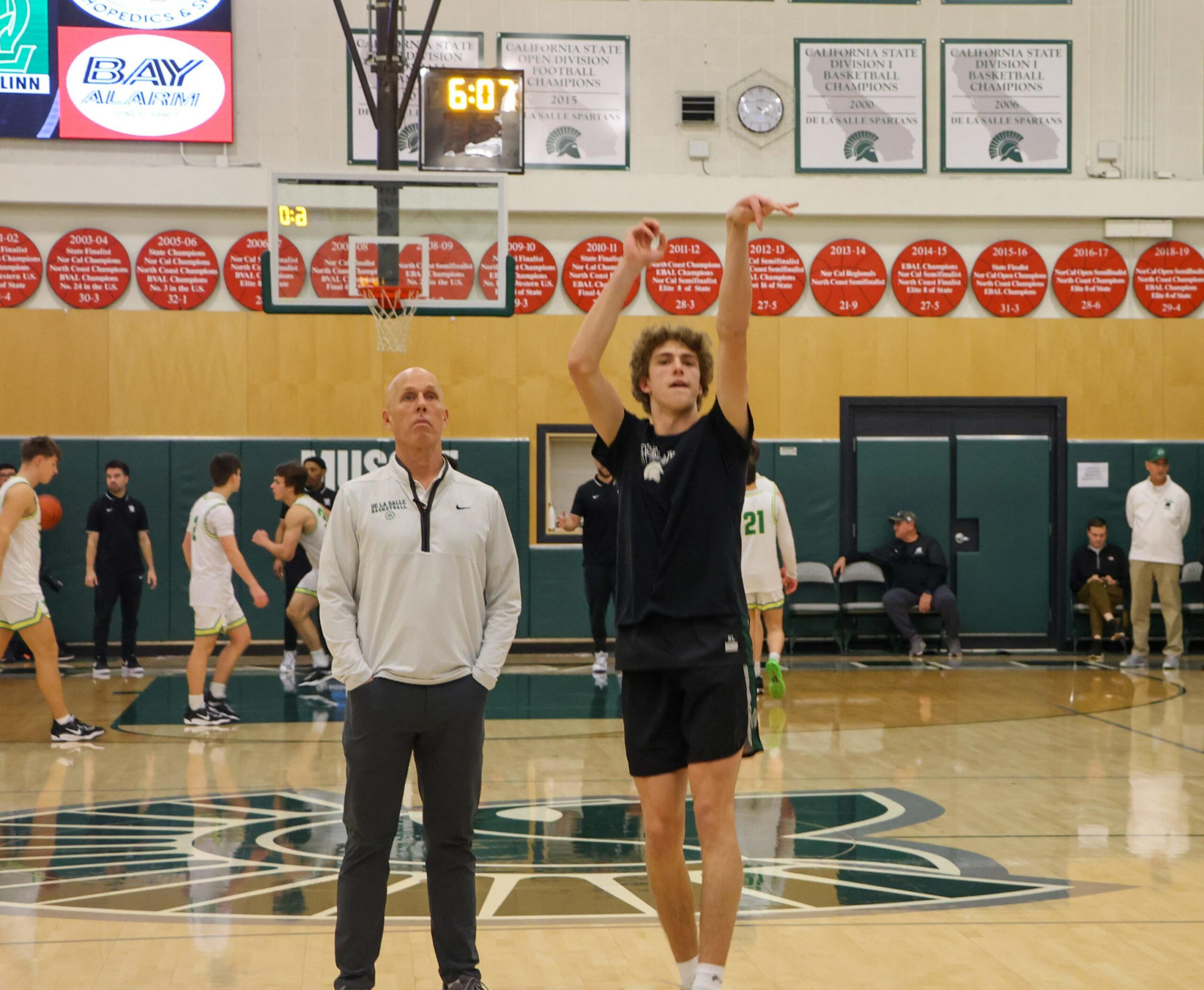 A young basketball player in black shoots a ball while a coach in white watches, with teammates warming up in the background on a gym court.