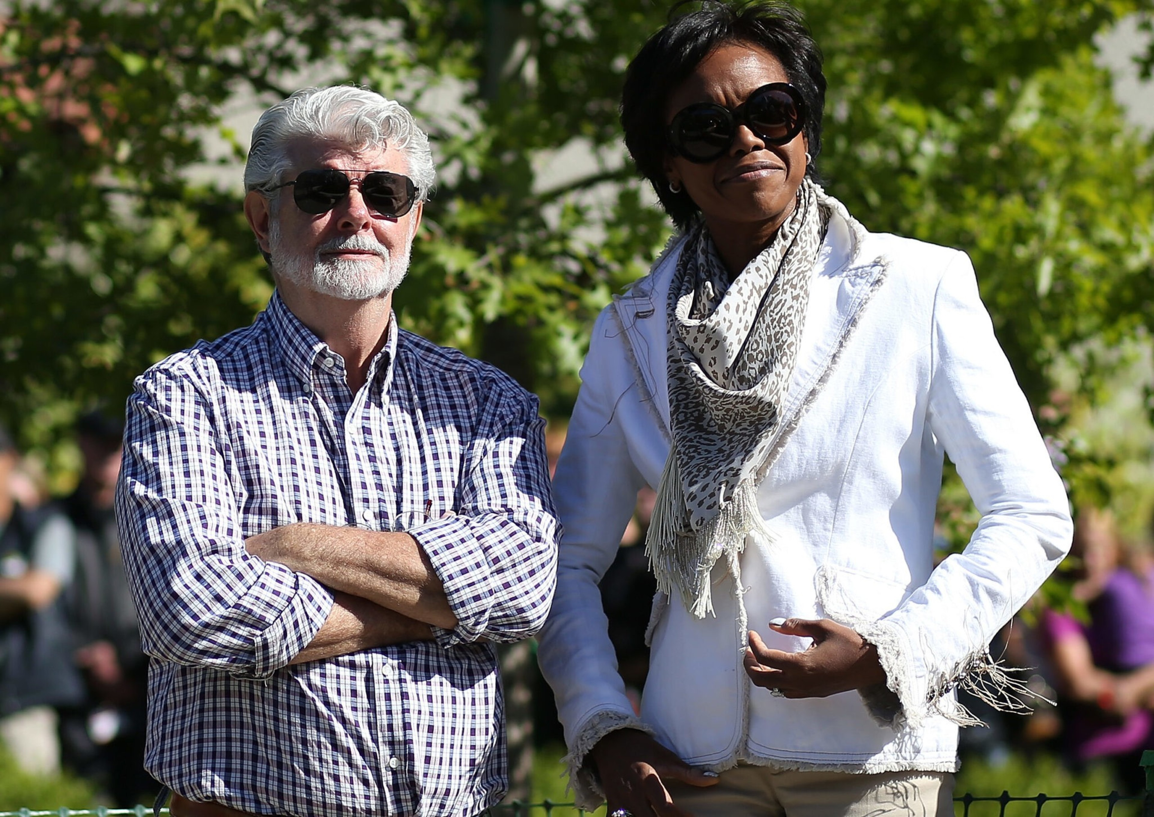 A man with white hair and beard and a woman with short hair wear sunglasses outdoors, standing side by side with a green leafy background.