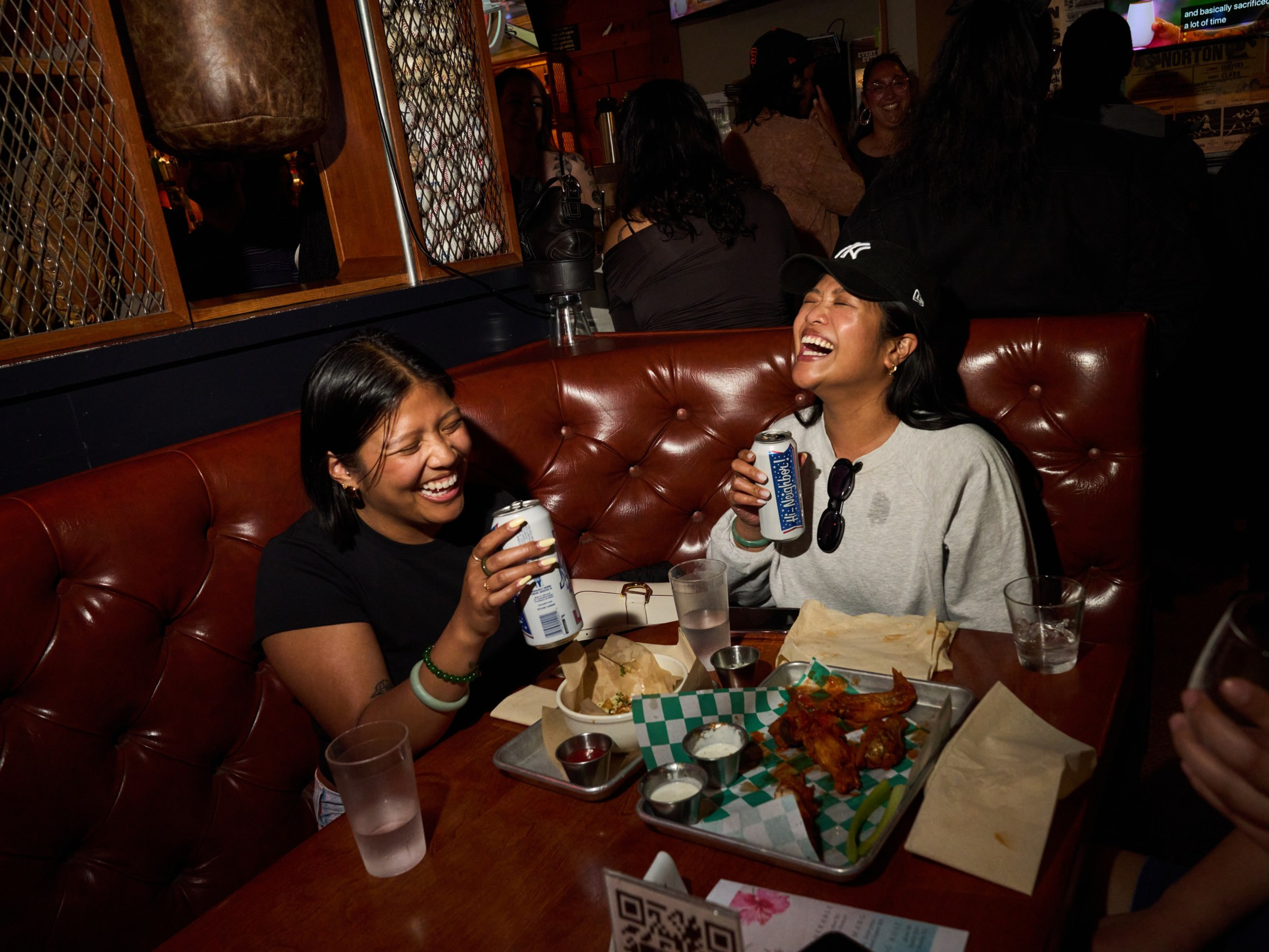 Two people are laughing and holding cans of beer in a dimly lit restaurant booth. There's a tray with food and empty glasses on the table.