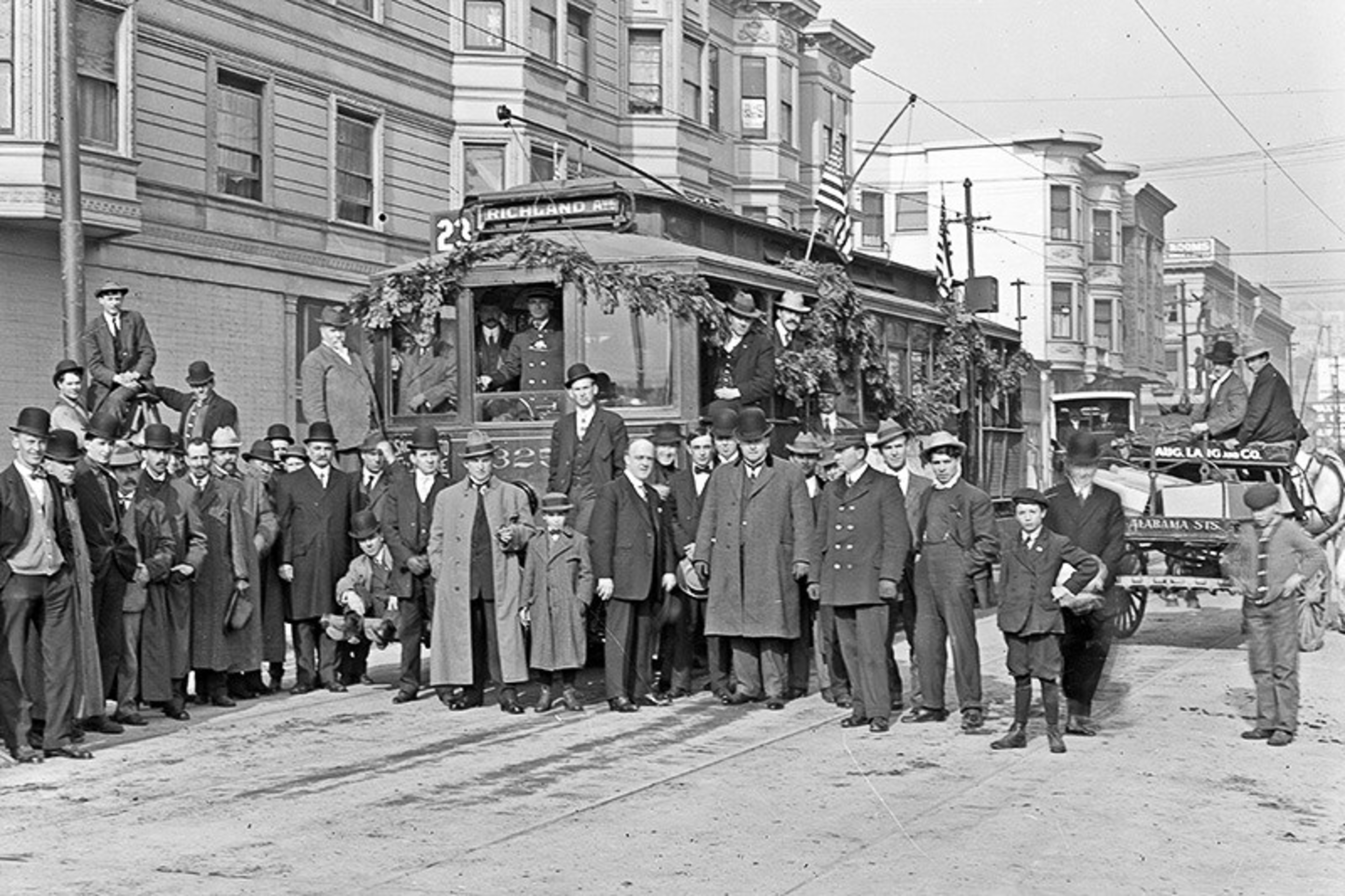 A group of men and boys in early 1900s clothing stand around a decorated streetcar labeled "23 Richland" on a city street with buildings in the background.