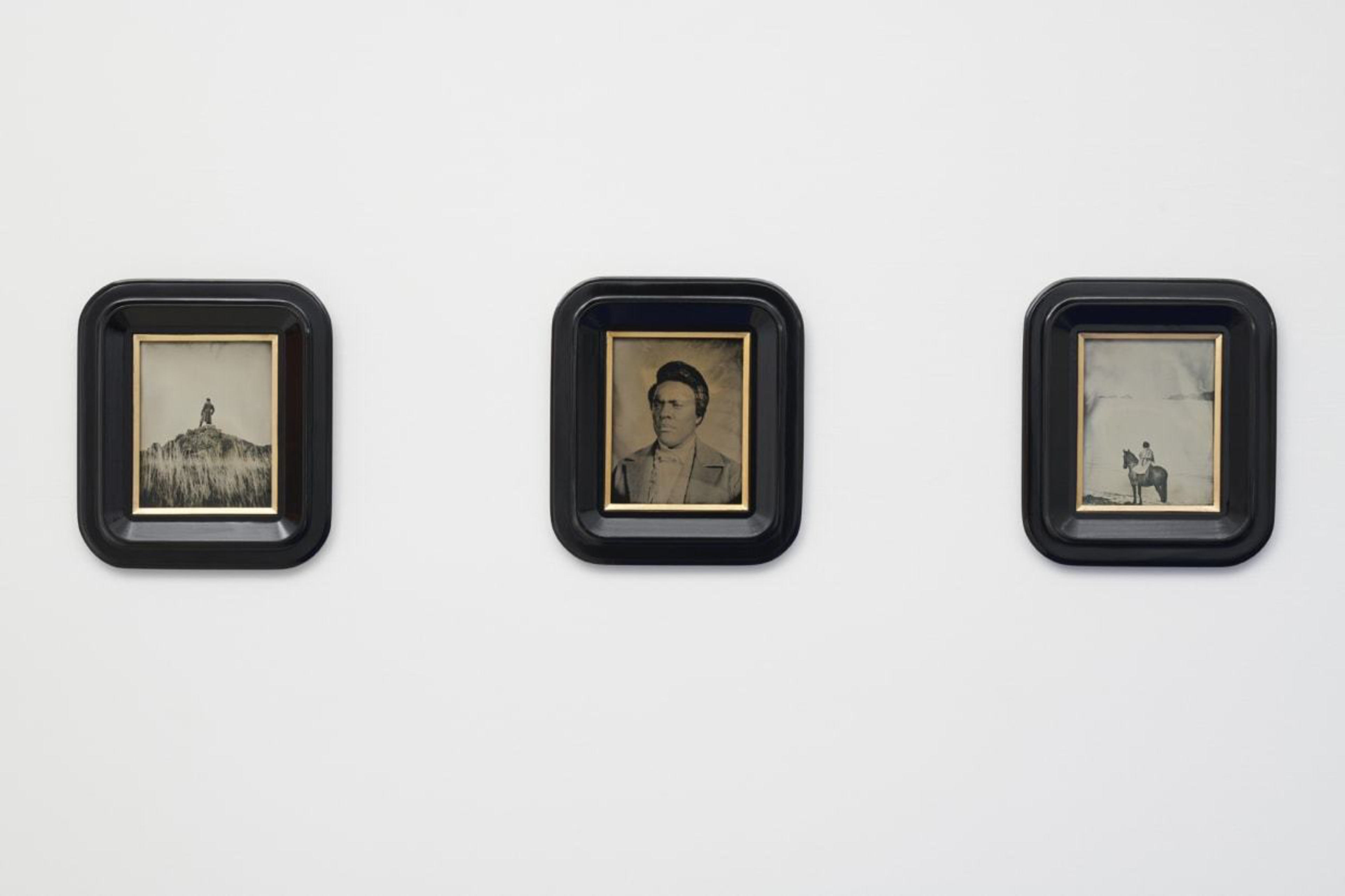 Three small black-framed vintage photographs hang on a white wall, featuring a person on a hill, a portrait, and a person on a horse.