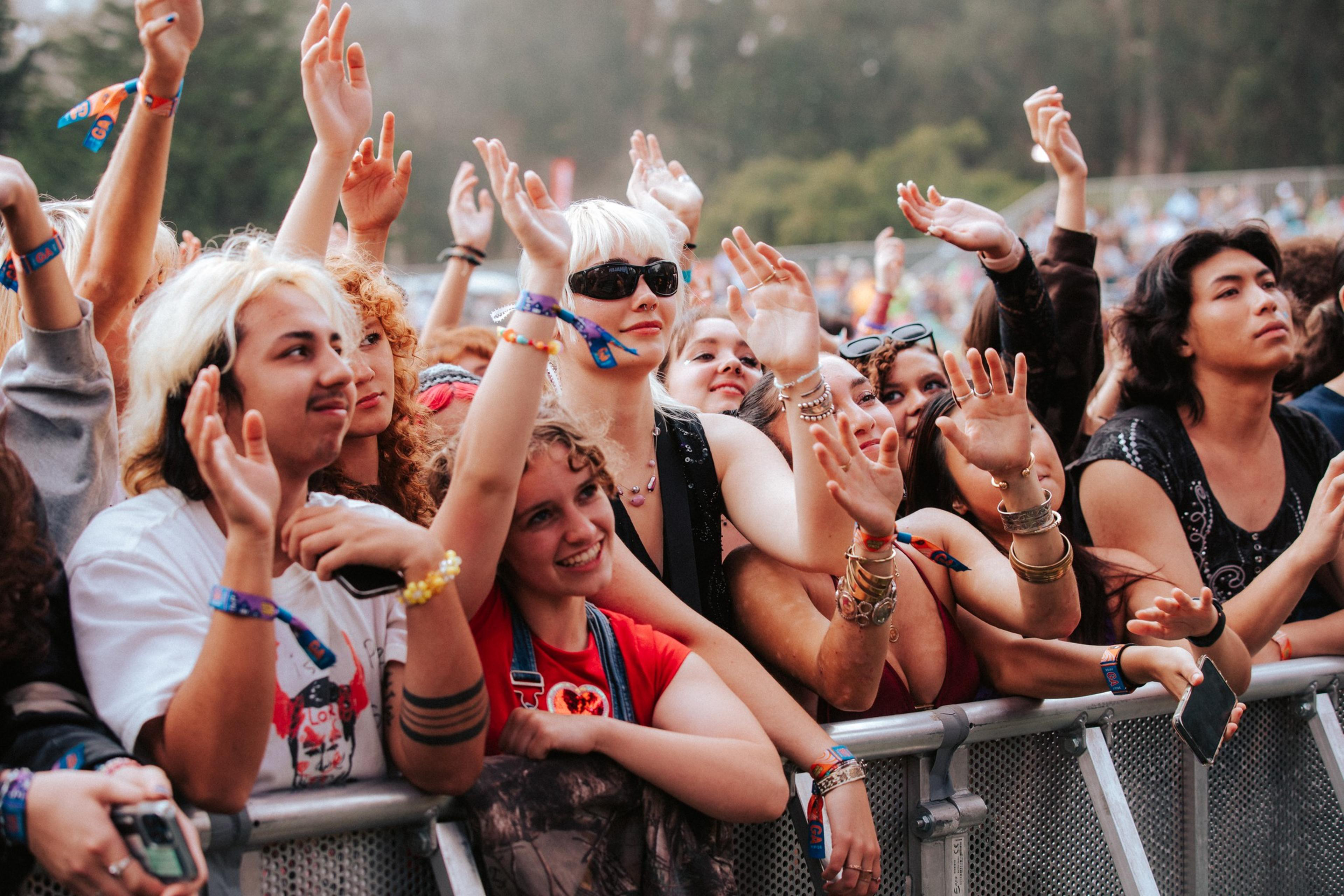A diverse group of people at a concert or event eagerly reach toward the stage. They are smiling, adorned with wristbands, and standing behind a metal barrier.