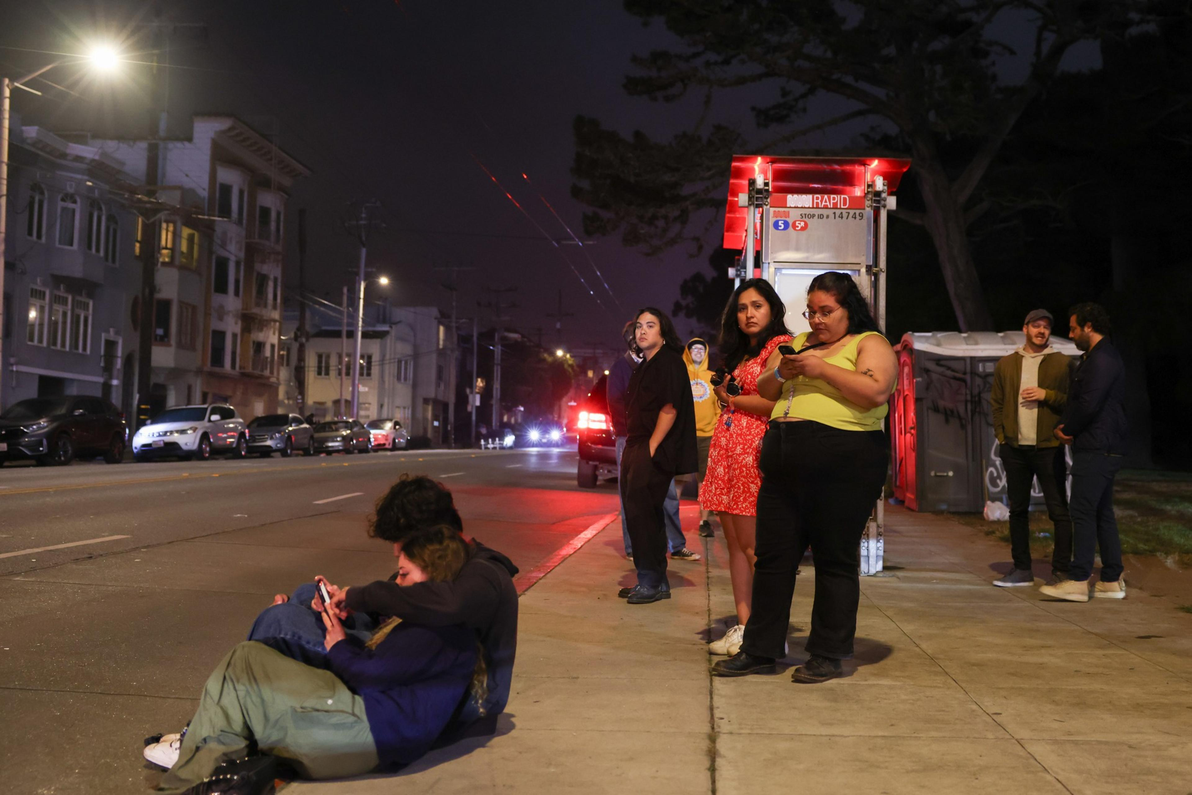 Several people wait at a bus stop on a city sidewalk at night, with some looking at their phones and others standing or sitting near the curb.