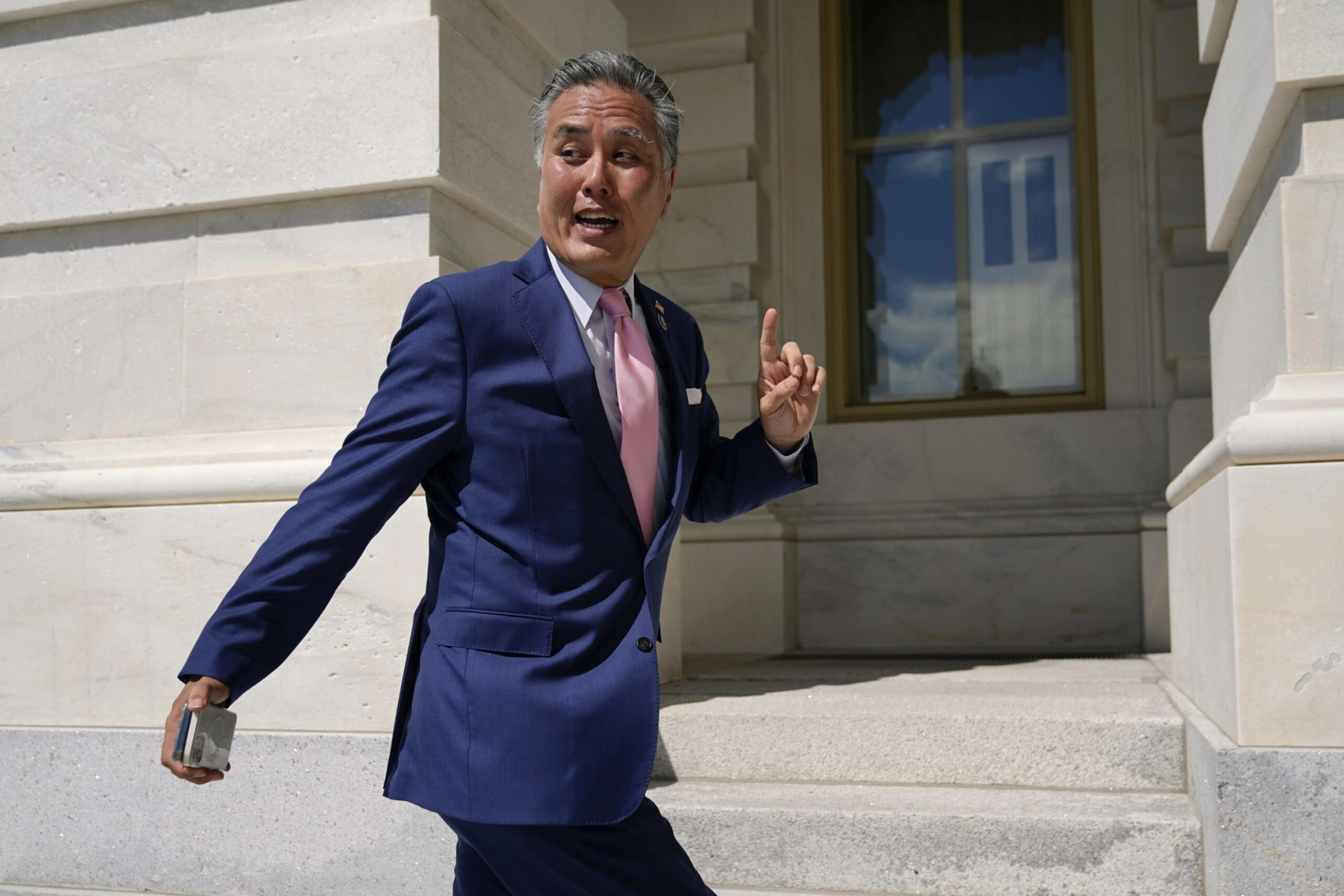 A man in a navy suit and pink tie walks briskly near a stone building, holding a phone in one hand and raising a finger with the other.