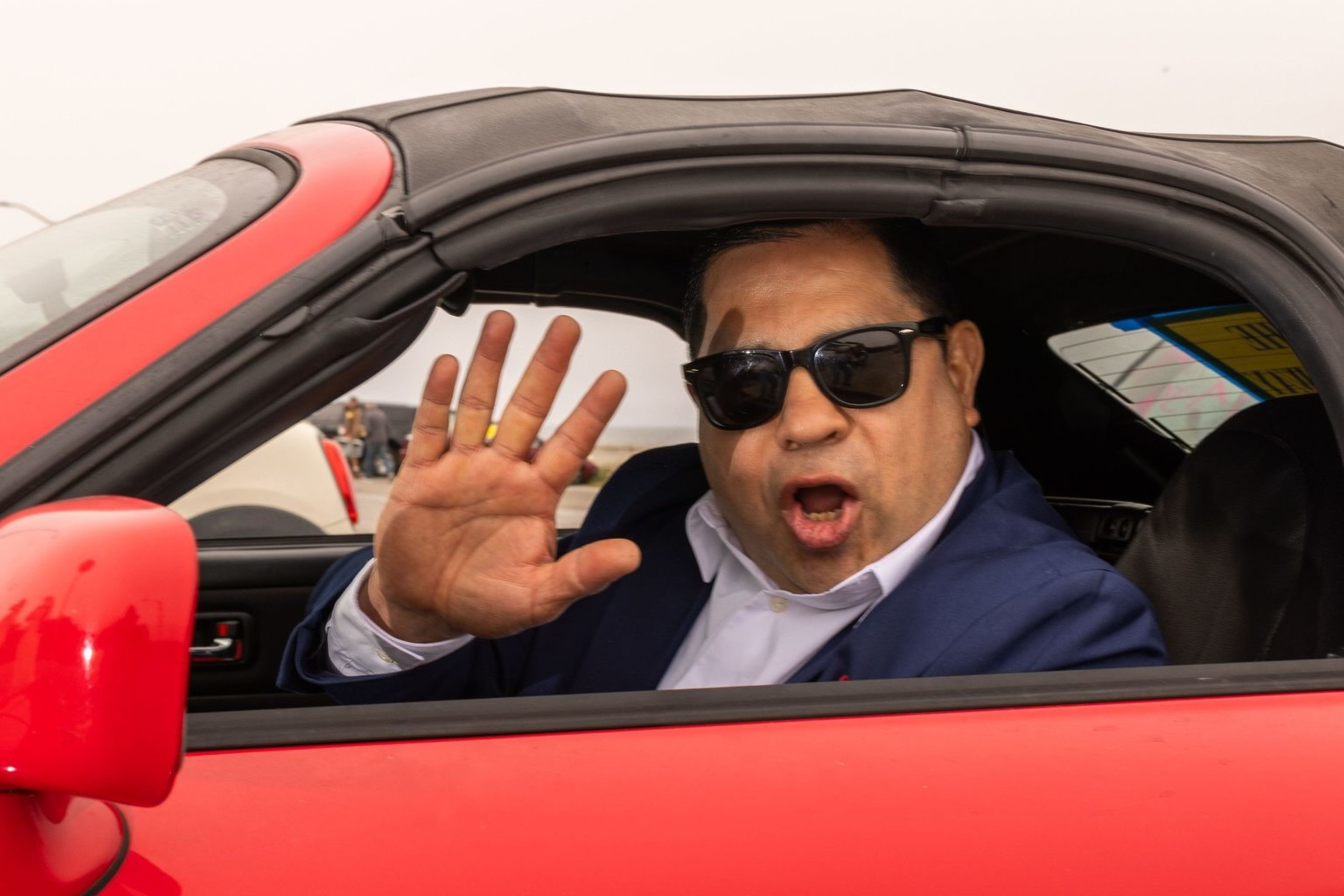 A man in sunglasses and a suit waves and speaks from a red convertible with a sign reading "OPEN THE GREAT HIGHWAY" in English and Chinese taped to the door.