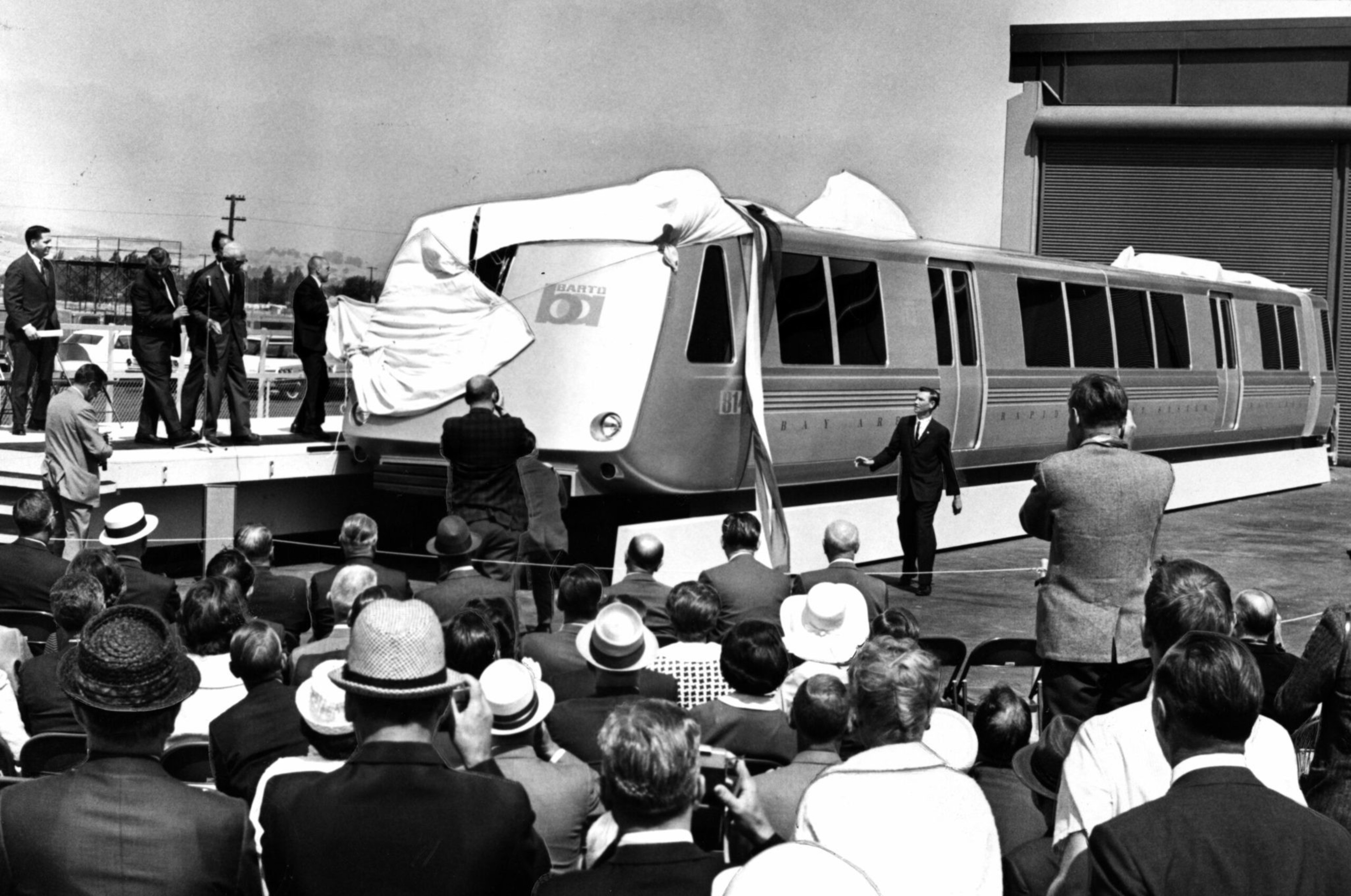 A crowd watches as a sleek, futuristic train car is unveiled on a platform in a black-and-white photo, with men in suits nearby.