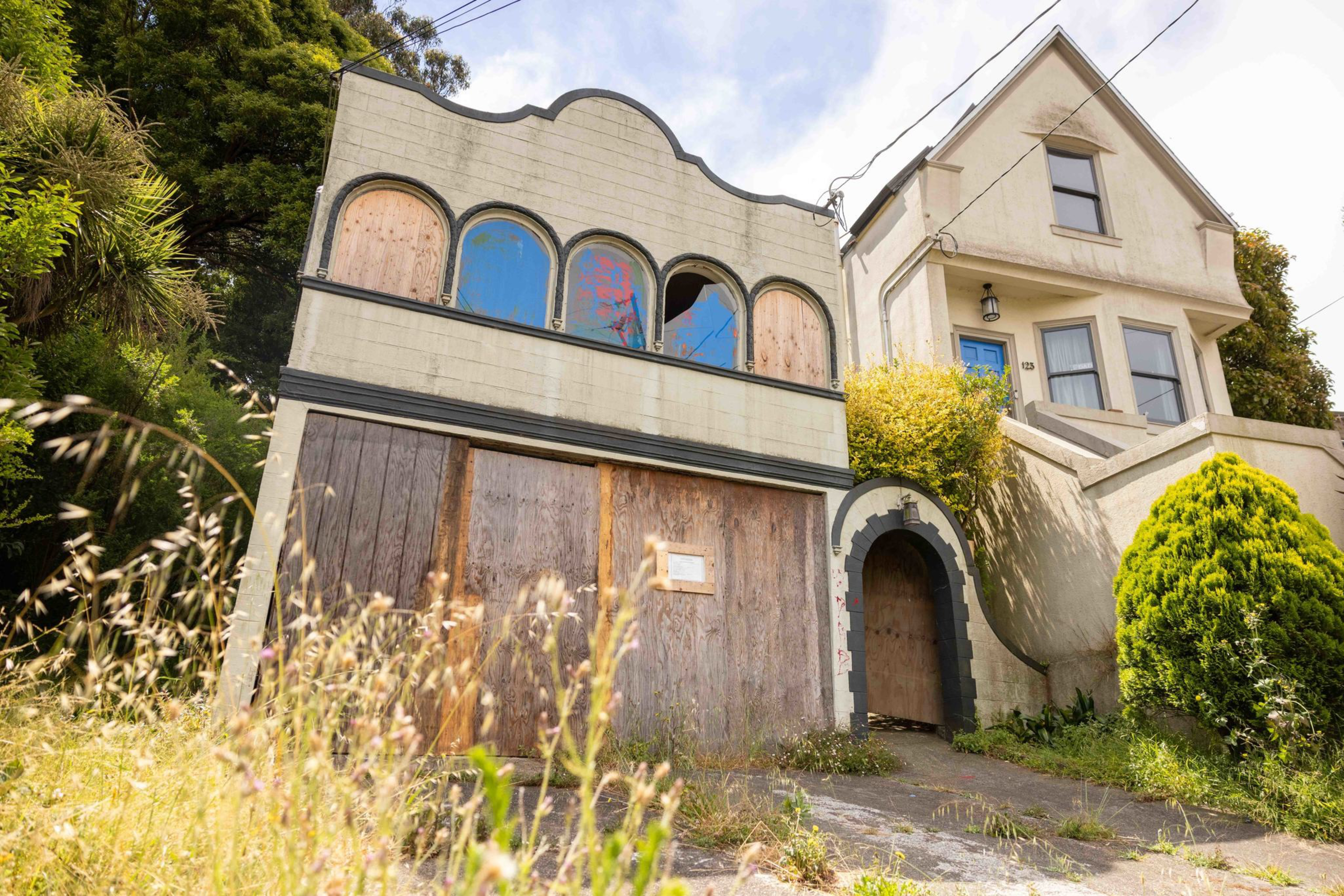 A two-story house with boarded-up windows and doors sits next to a maintained beige house with a blue door, surrounded by overgrown grass and bushes.