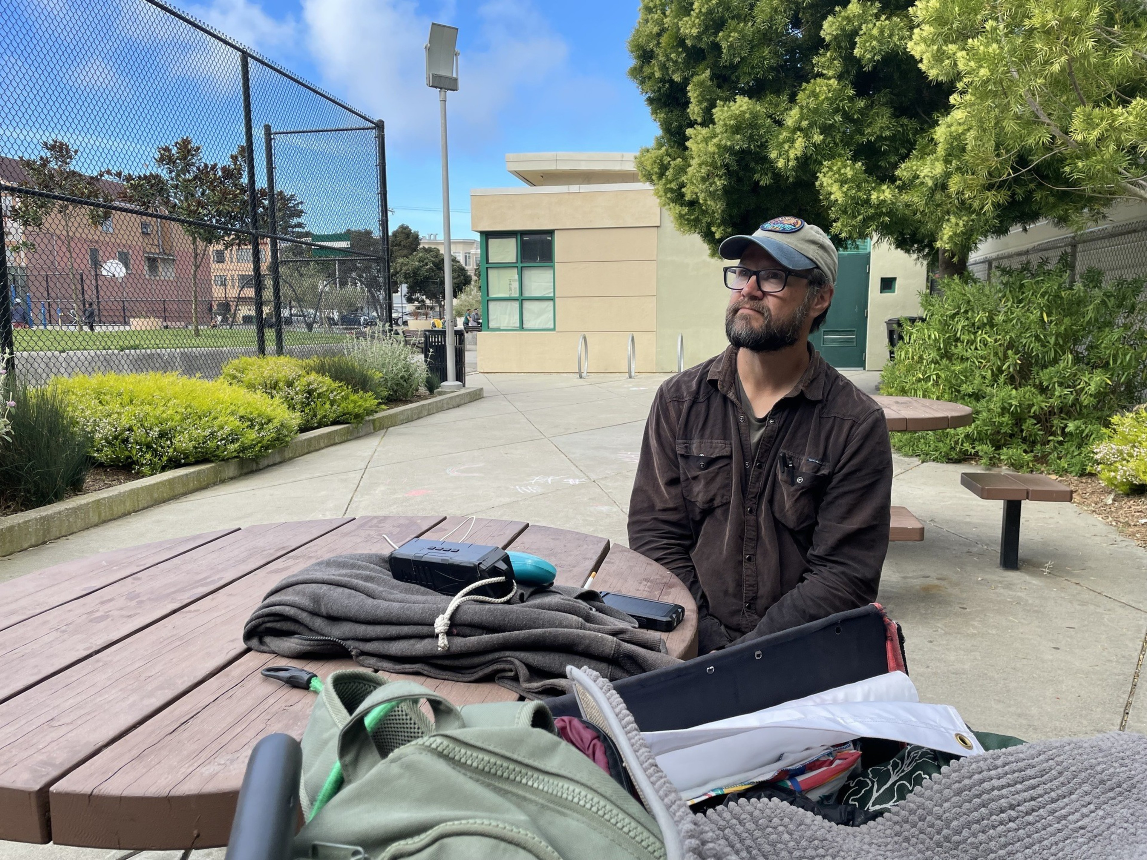 A man sits at a table in a park with belongings in a wagon nearby.