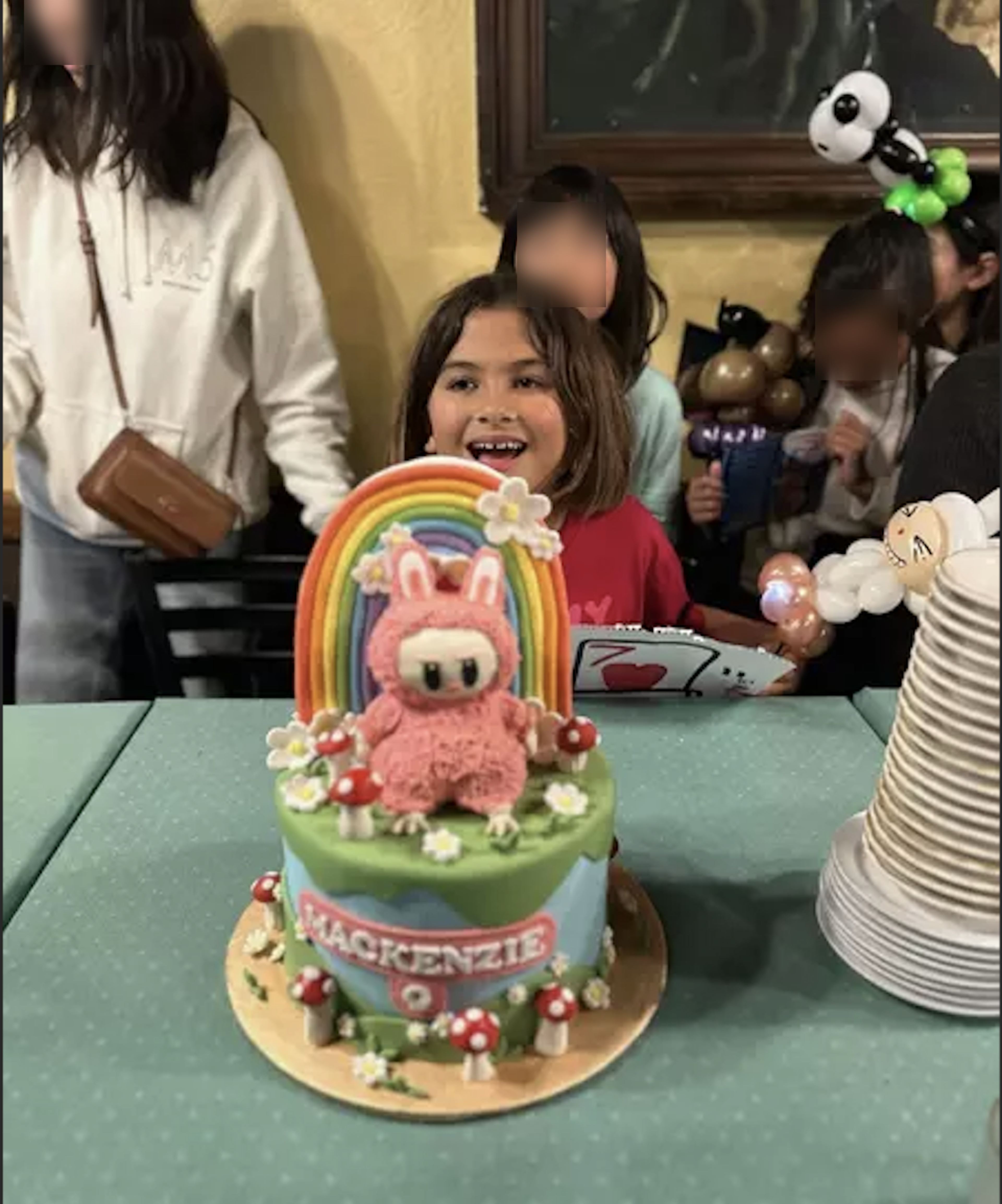 A colorful birthday cake with a rainbow, a pink bunny figure, mushrooms, and "Mackenzie" written in front, with a smiling child and party guests in the background.