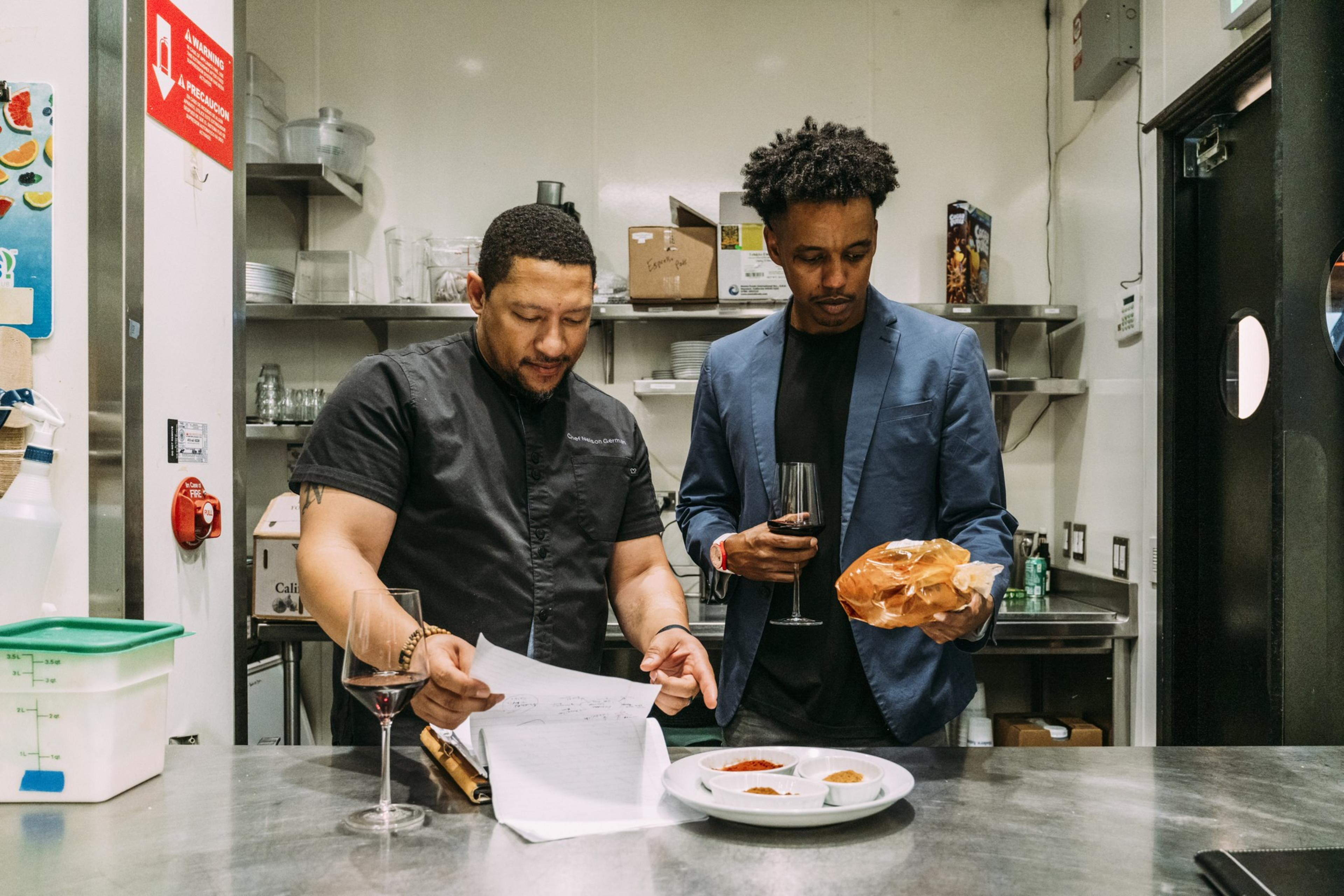 Two men stand in a kitchen, one holding papers and the other holding bread and a wine glass. Spices in small bowls are on the counter.