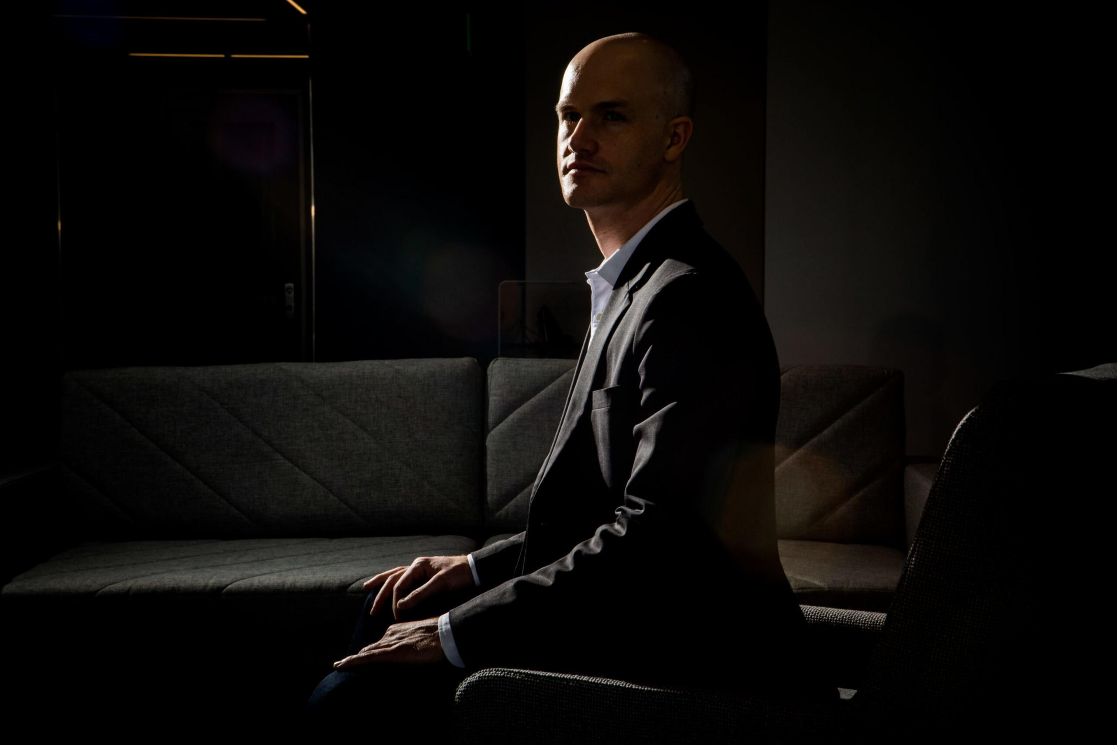 A person in a dark blazer sits in a dimly lit room on a gray, textured couch. The lighting creates a dramatic effect, highlighting their profile.