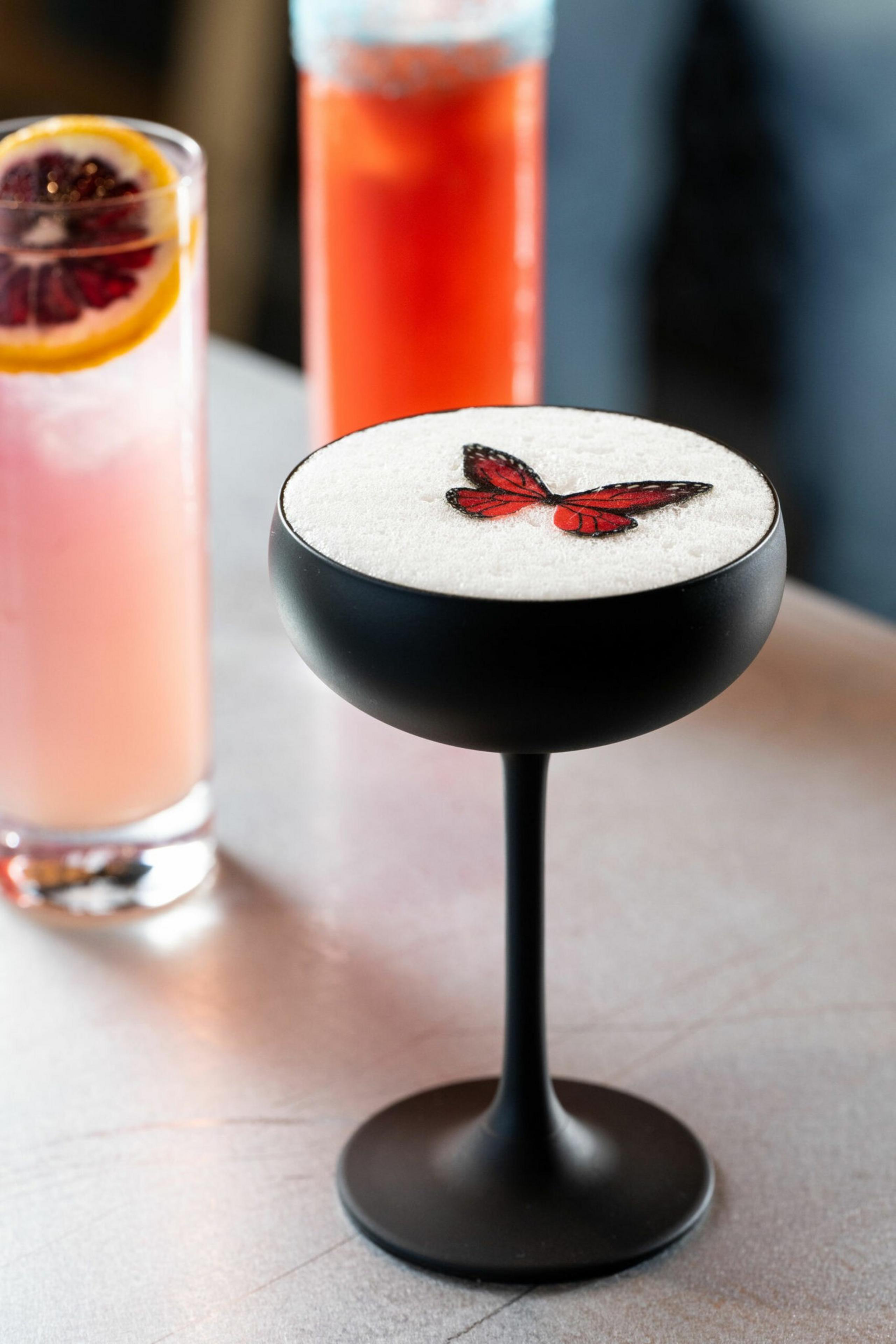 A black coupe glass holds a frothy white cocktail topped with a red butterfly decoration, with two colorful drinks blurred in the background.