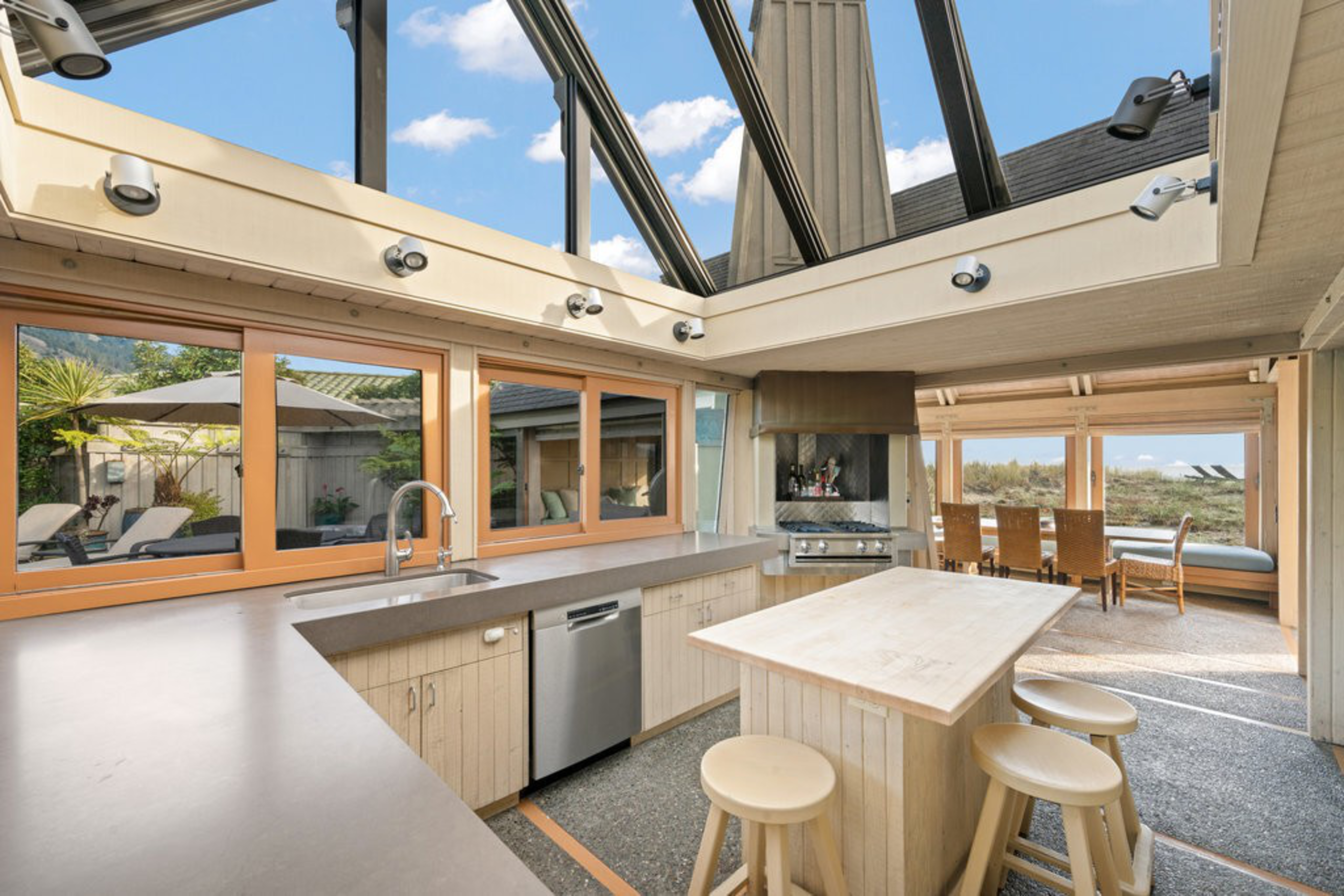 A modern kitchen with beige countertops, wooden stools around a central island, large windows, a skylight ceiling, and a dining area in the background.