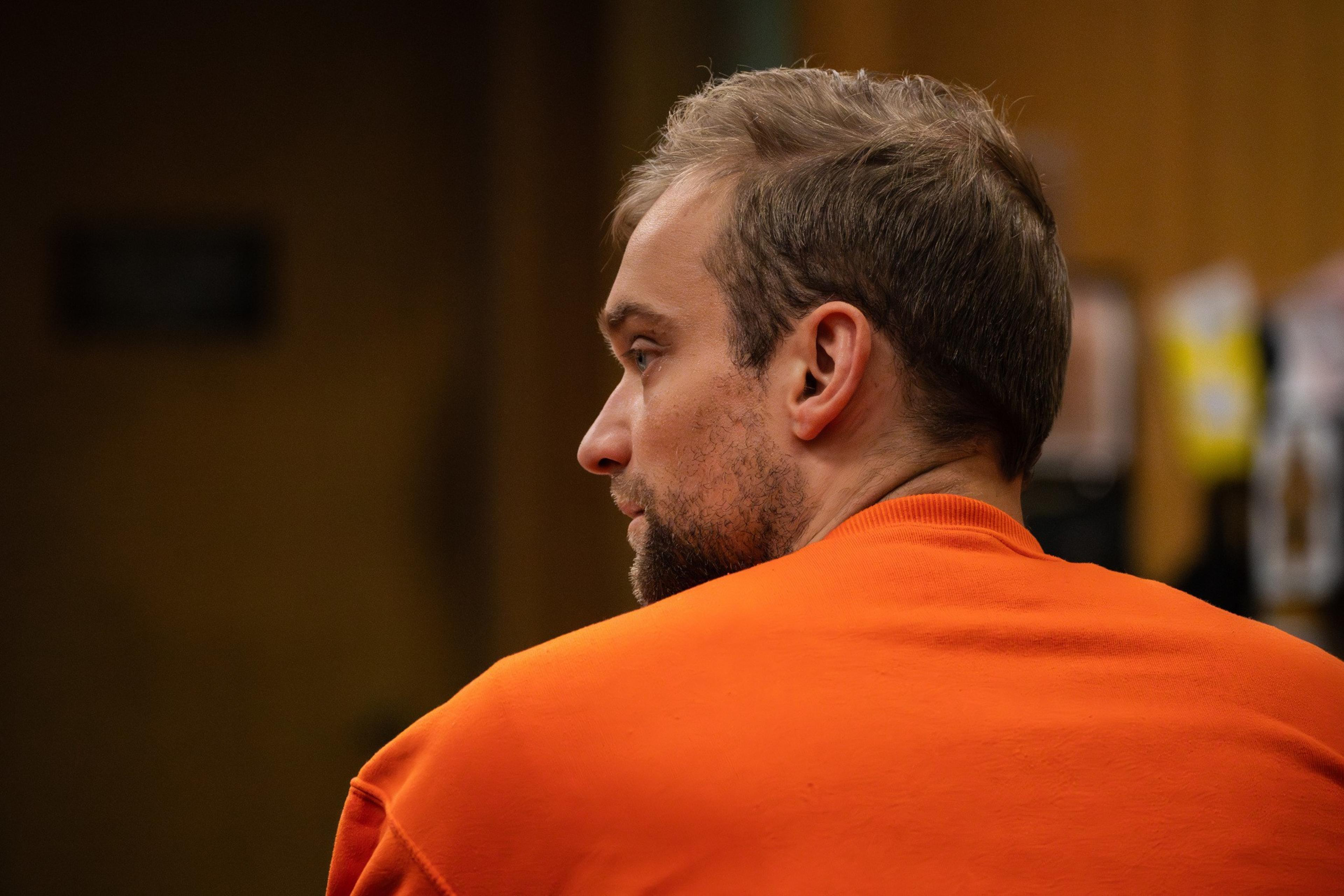 A person in an orange jail outfit sits in court and looks to the side.