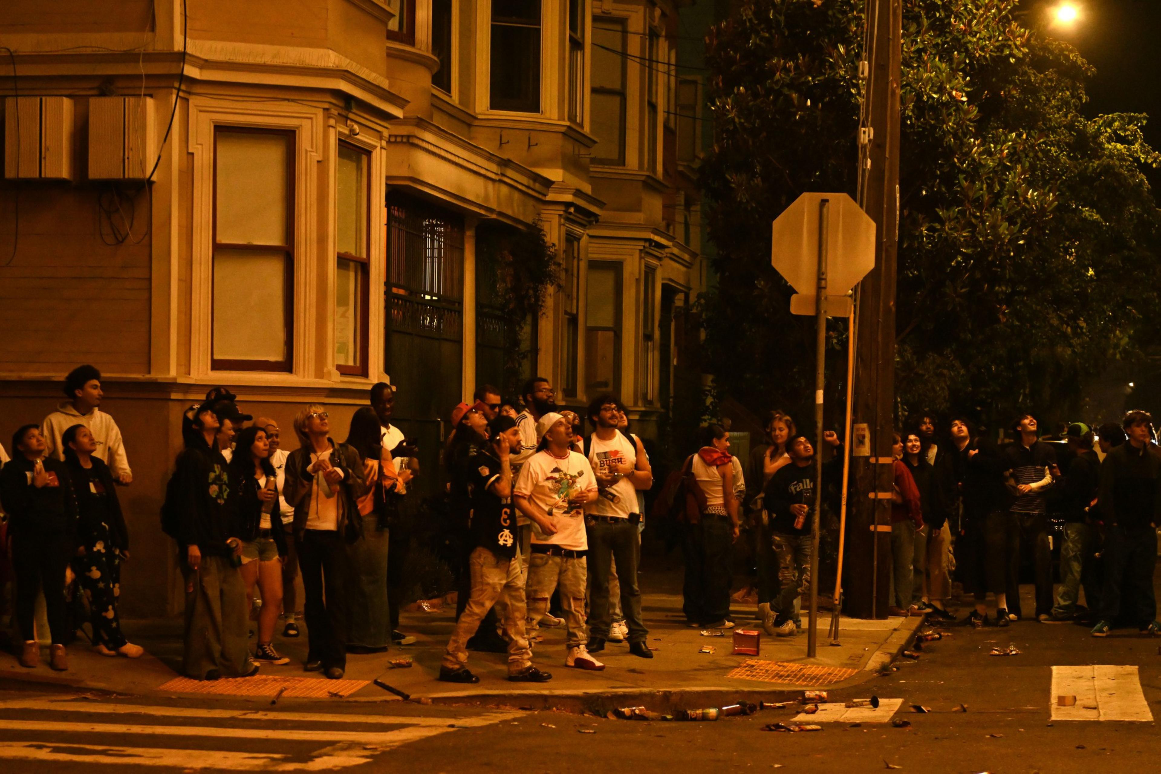 A group of people stand on a city street at night, looking upwards at fireworks in the night sky. They are gathered near a building with bay windows, and the ground is littered with trash.
