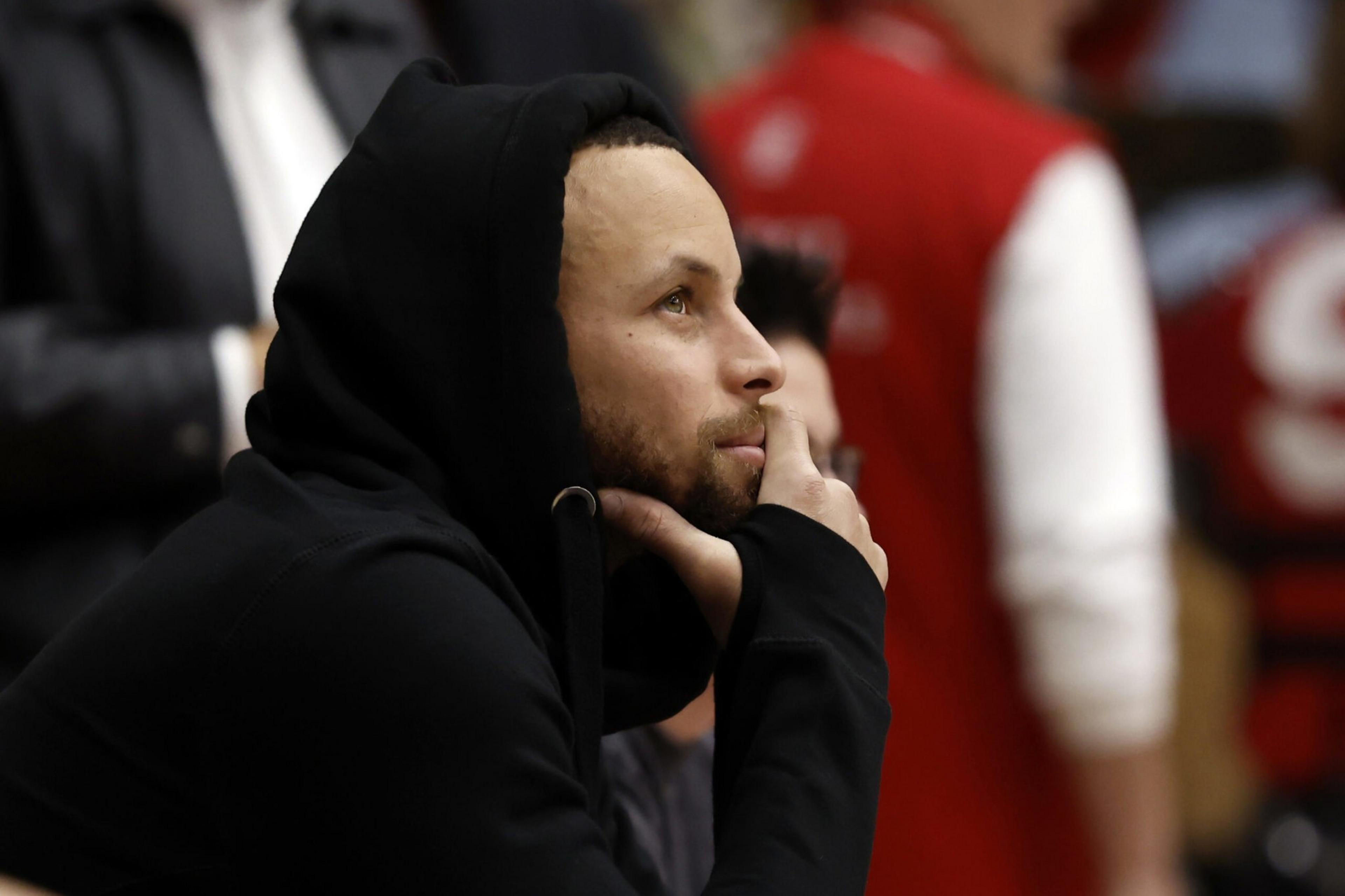 A person wearing a black hooded sweatshirt looks thoughtful, resting their chin on their hand. The background shows blurred figures and muted colors.