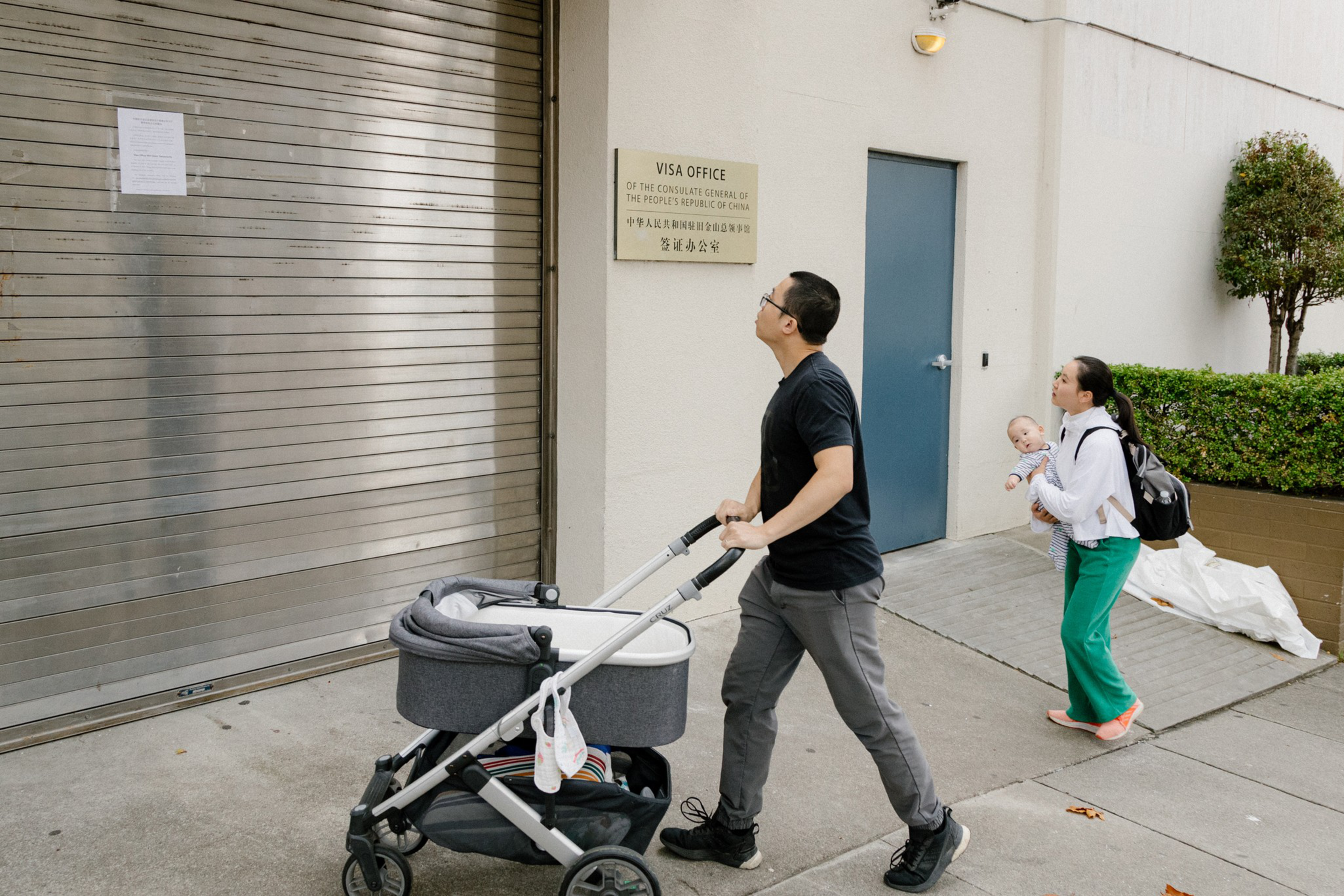 A man with a stroller and a woman carrying a baby look at a notice posted on a closed metal shutter outside a visa office.