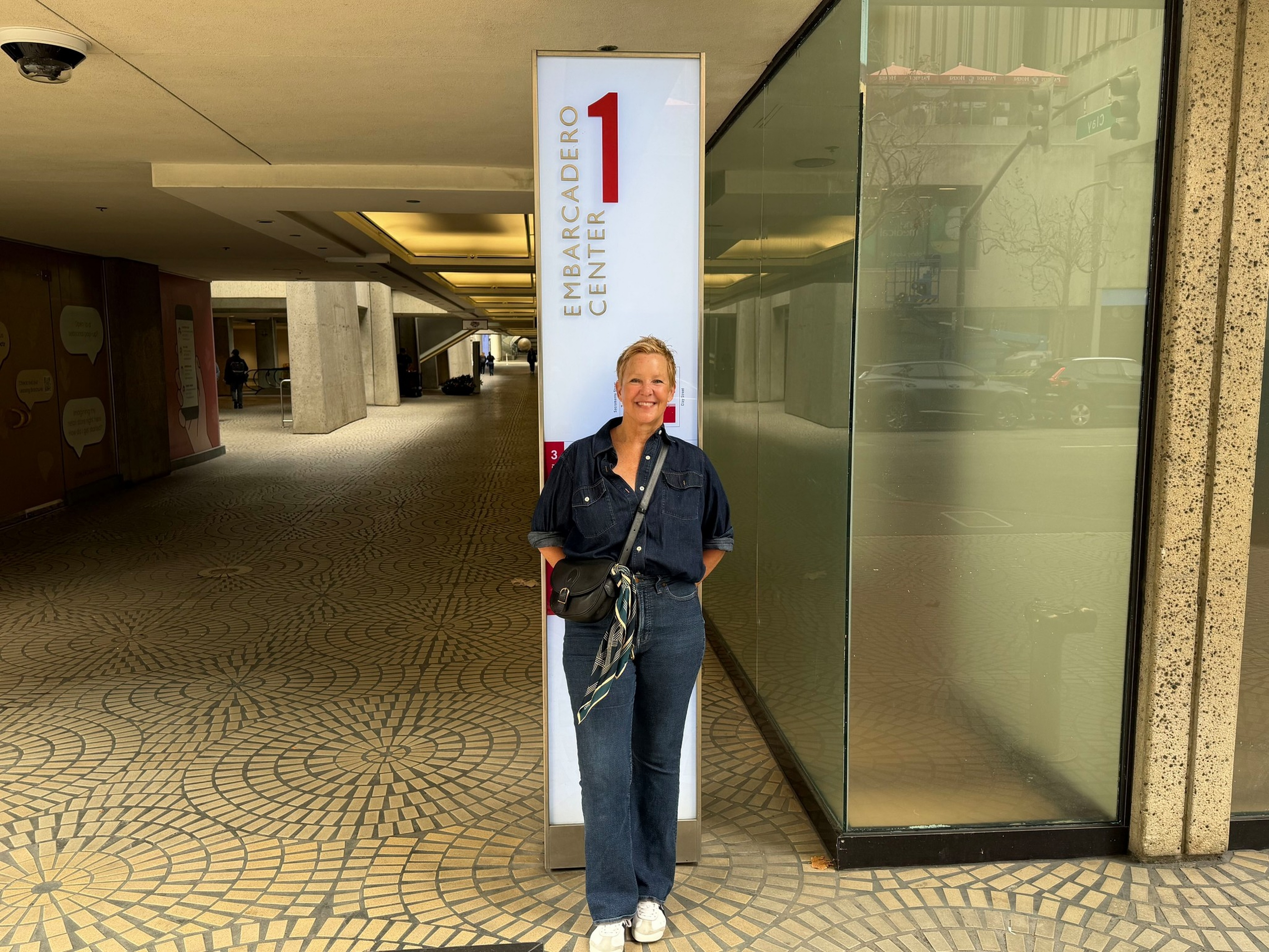 A woman in a dark denim outfit and white sneakers stands smiling in front of a tall sign that reads "Embarcadero Center 1" in a patterned tiled walkway area.