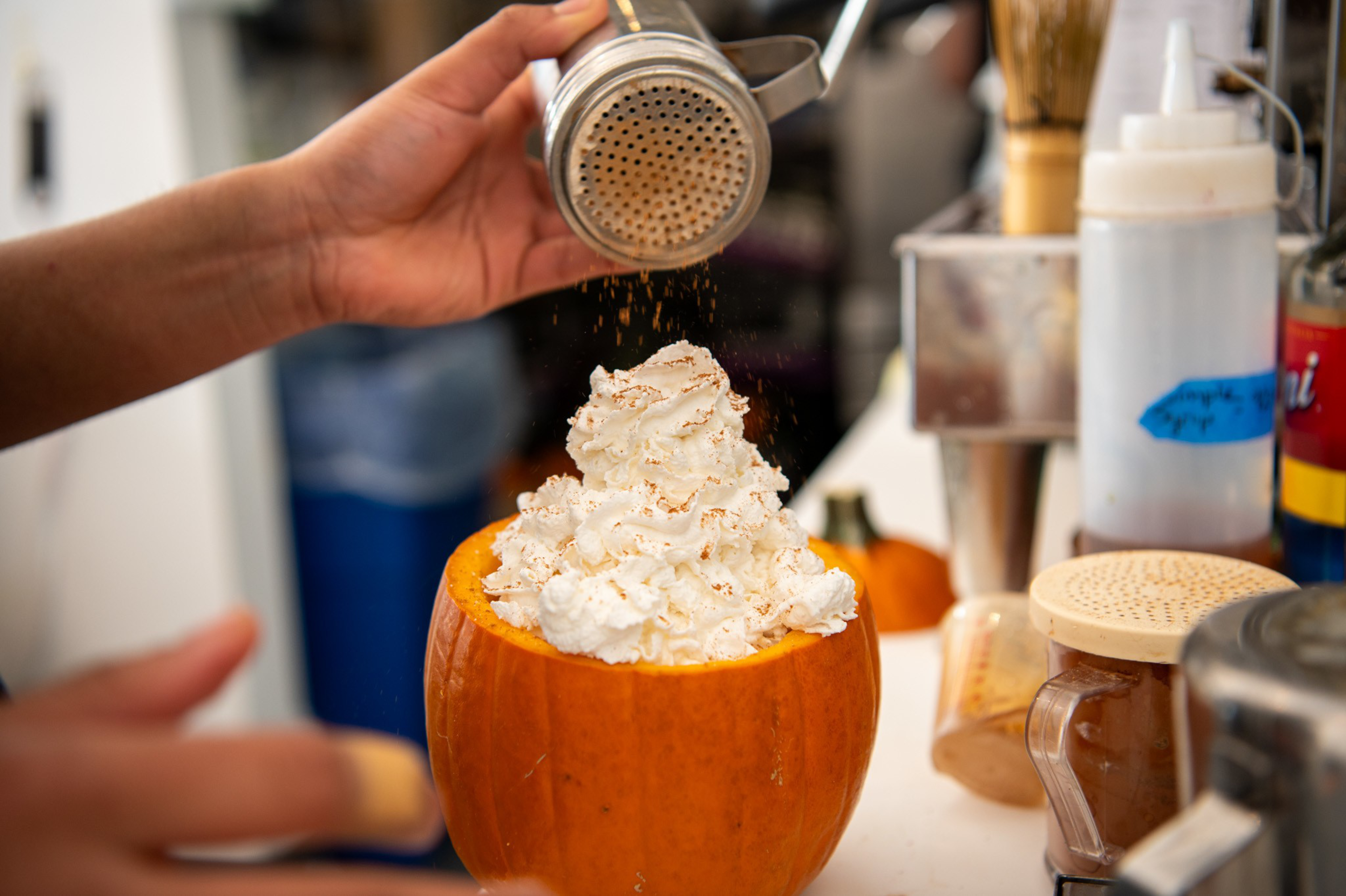 Whipped cream is piled high inside a hollowed-out pumpkin while a person sprinkles cinnamon or nutmeg on top using a metal shaker.