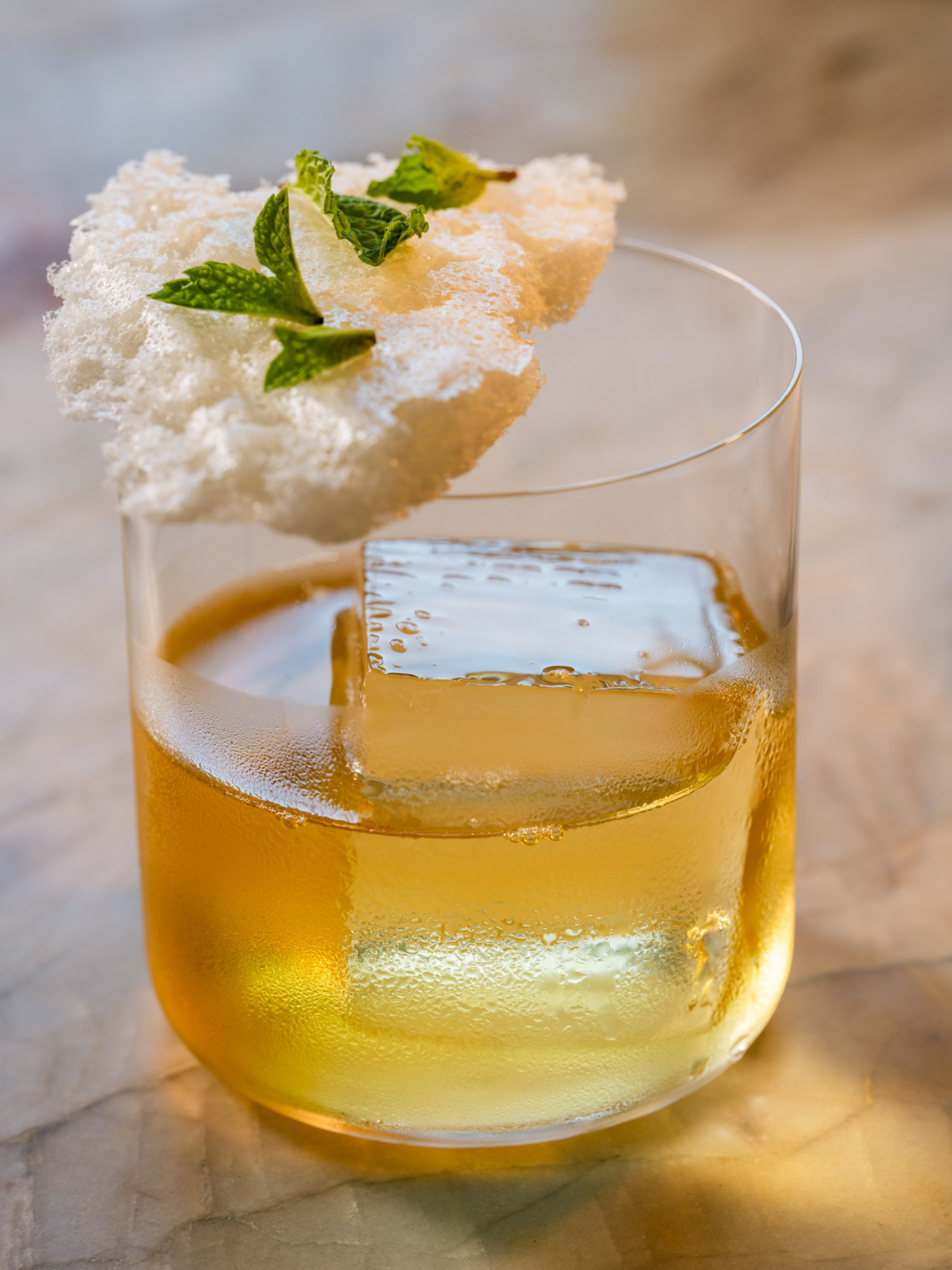 A glass contains a golden drink with a large clear ice cube, topped with a white crispy garnish and small green mint leaves.