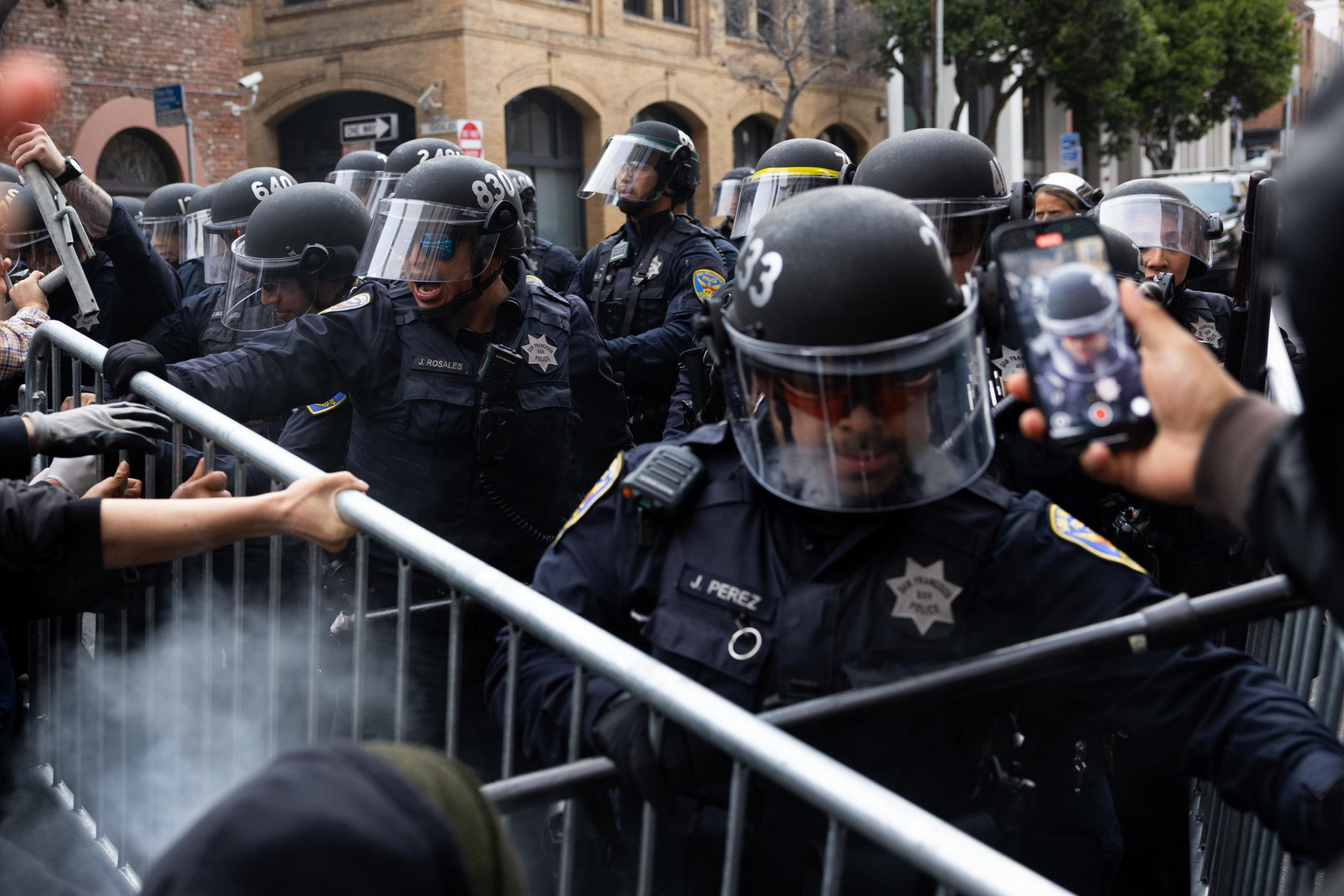 Police officers in riot gear push against metal barricades held by protesters, with some people capturing the scene on their phones.