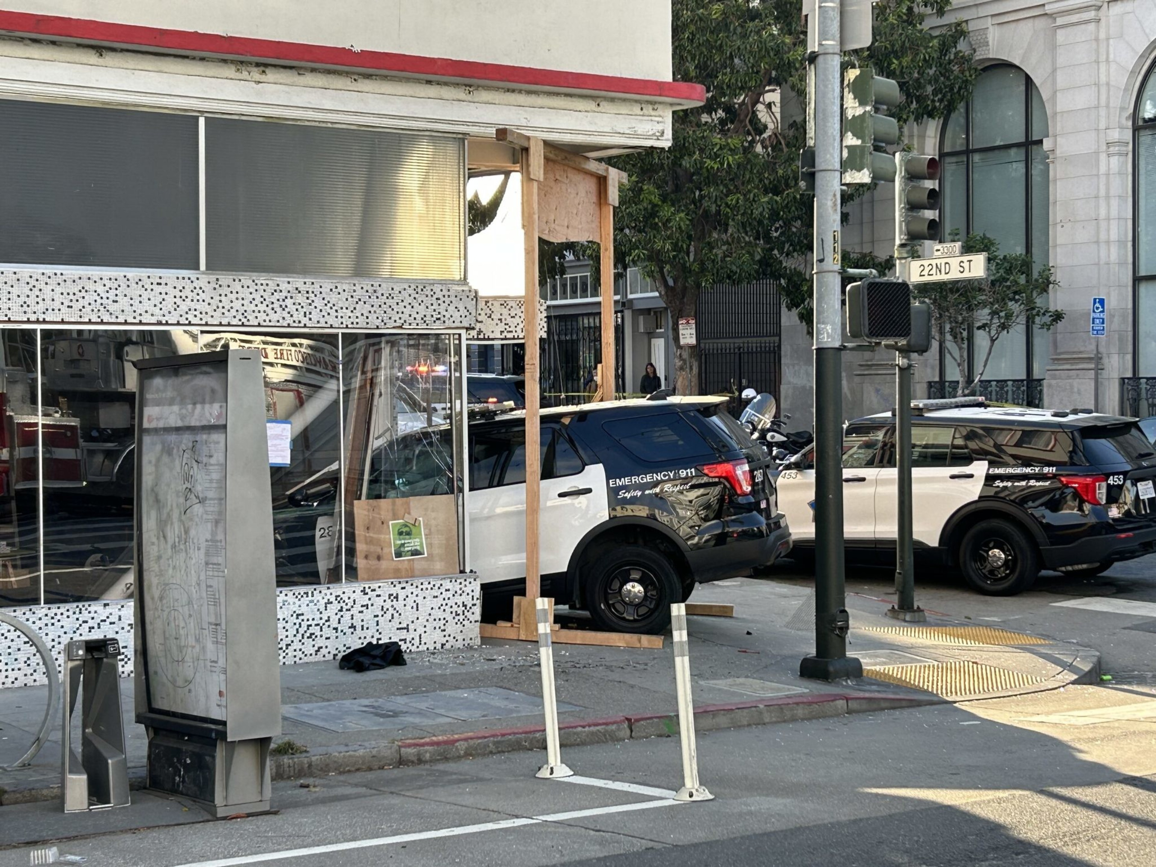 A damaged police SUV is crashed against a building near a street corner with traffic lights and another police vehicle nearby.
