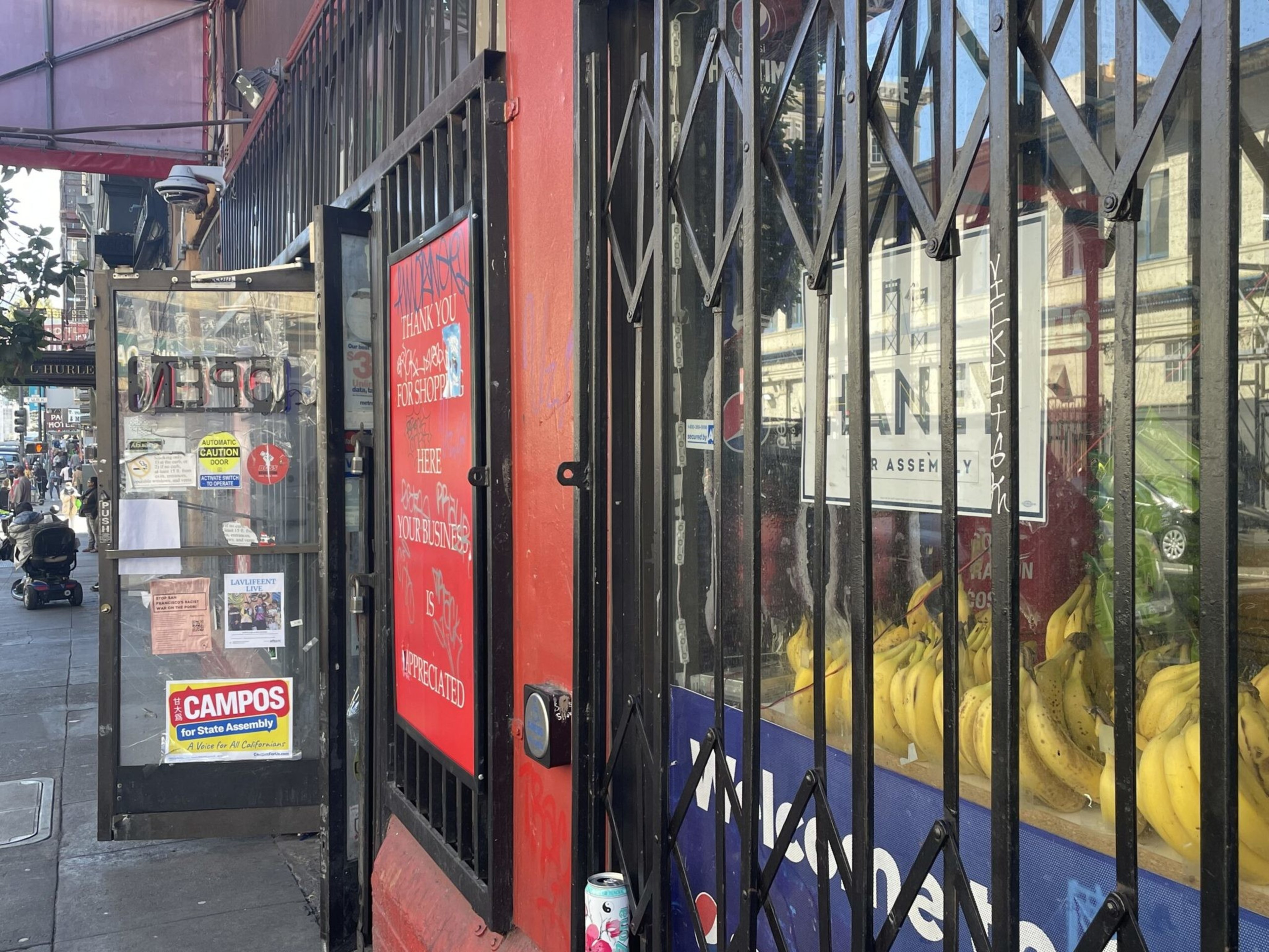 Bananas are displayed behind metal bars next to a red wall with a sign, and a glass door with various stickers is open on a busy city sidewalk.