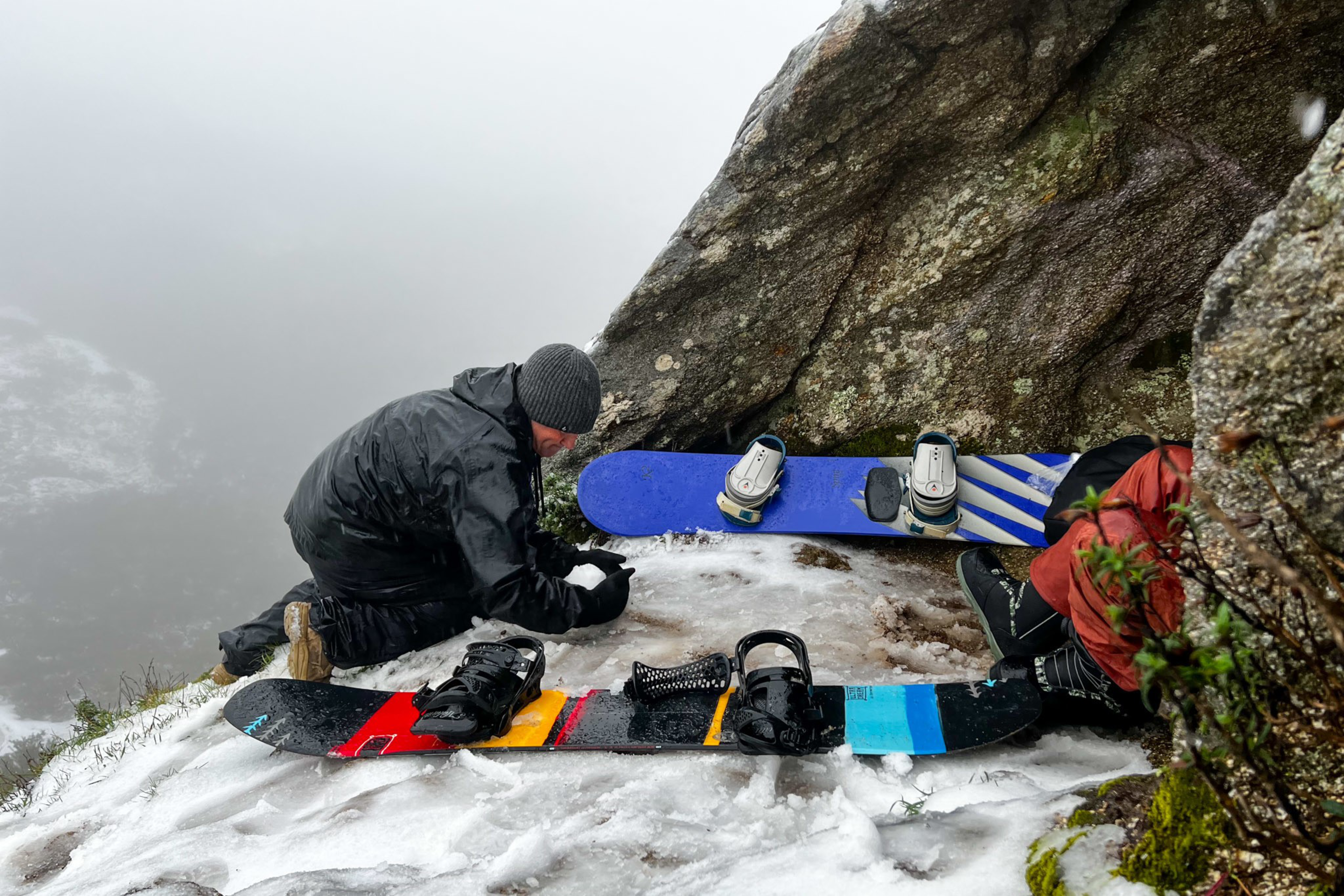 Two people dressed in winter gear rest on a snowy ledge beside large rocks, with two snowboards placed on the icy ground nearby.