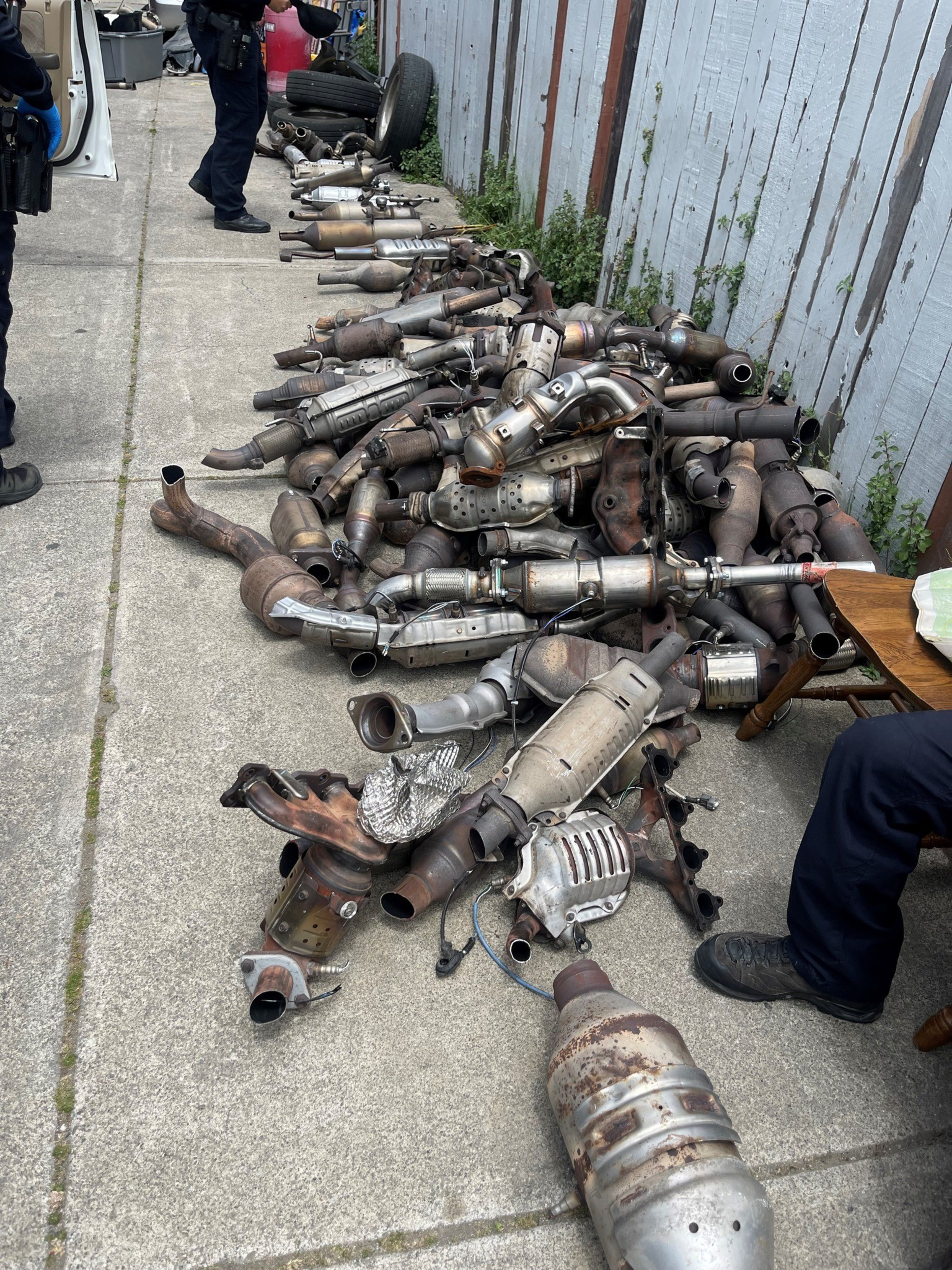 A large pile of catalytic converters found in raids are strewn across a sidewalk against a wooden fence.