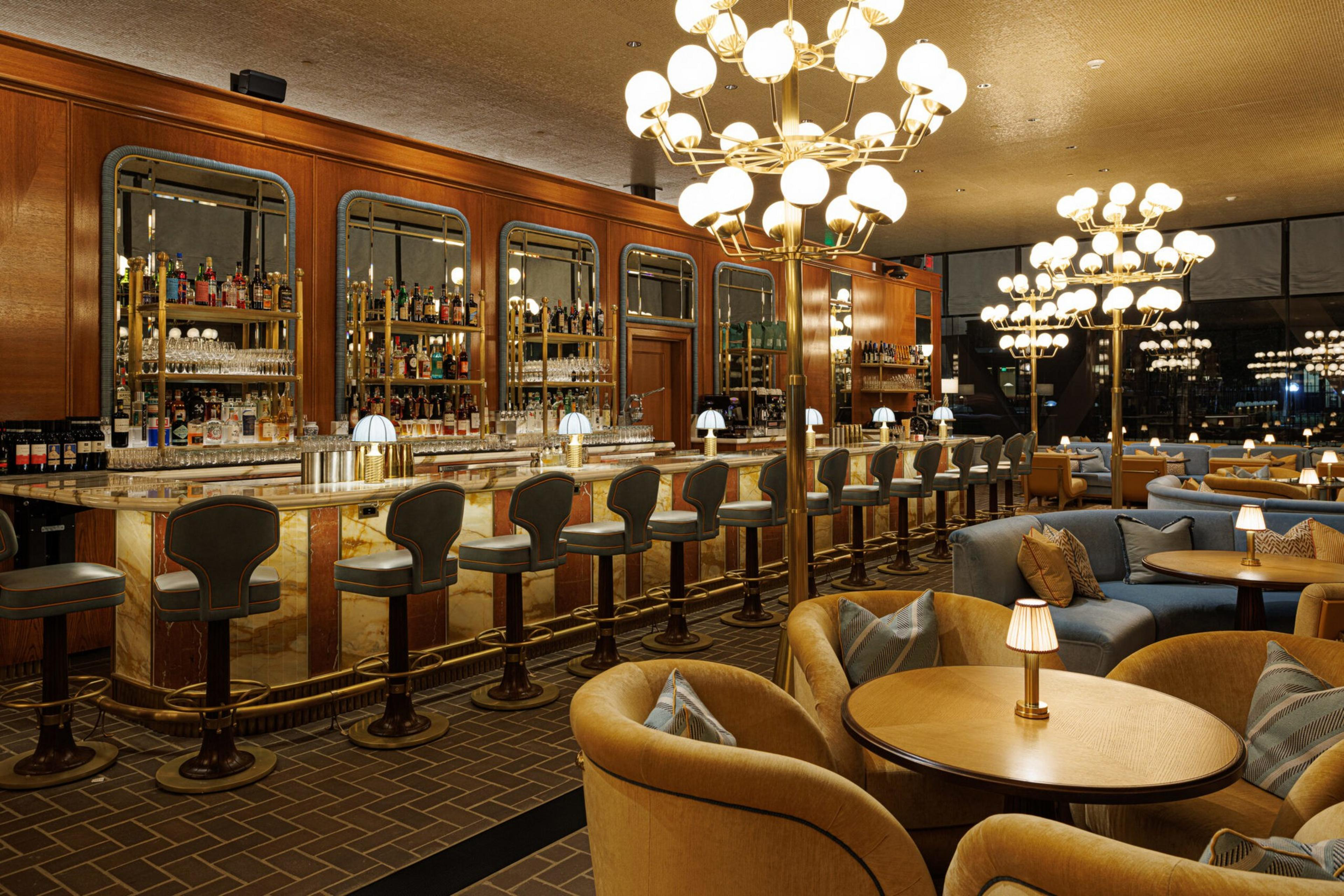 A luxurious bar with high stools, round tables with small lamps, cushioned chairs, and elegant globe chandeliers hanging from the ceiling.
