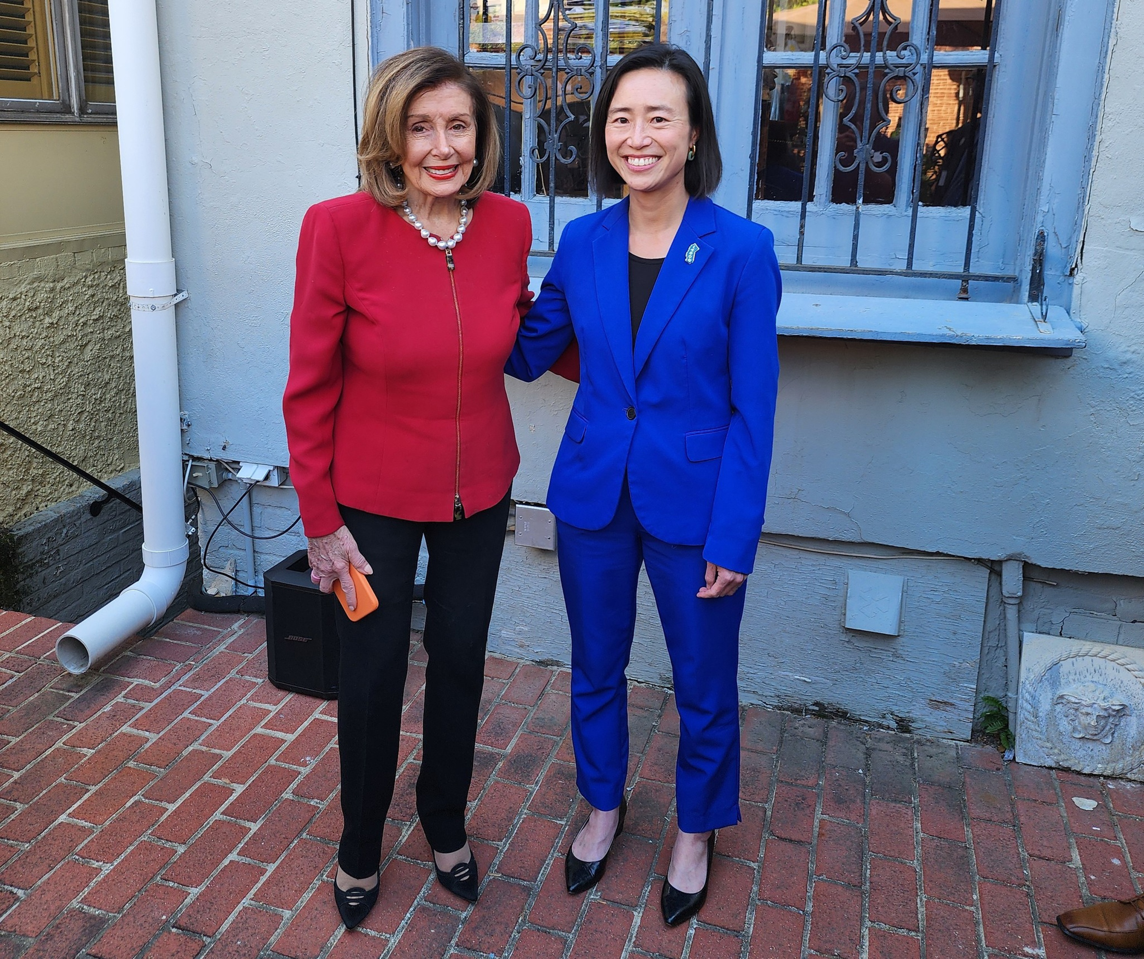 Two women stand on a brick patio; one wears a red jacket and black pants, and the other wears a royal blue suit, both smiling and posing together.