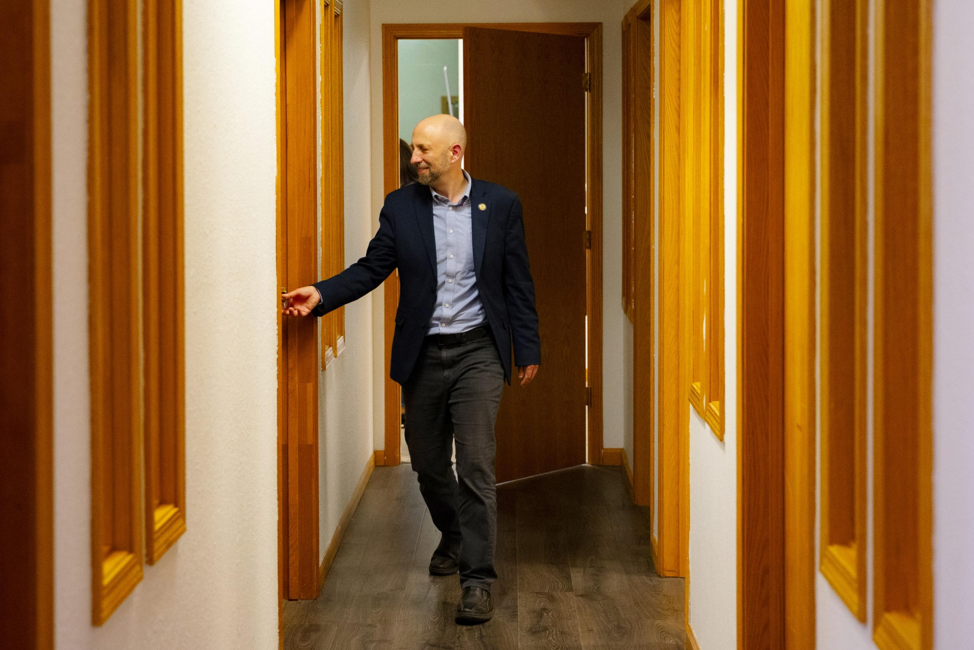 A bald man in a blazer and shirt walks down a wood-trimmed hallway, reaching out to touch the light switch on the wall.